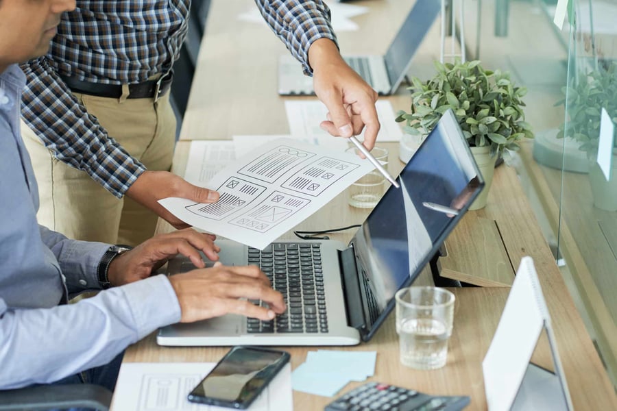 Two professionals reviewing website wireframes on paper and a laptop, discussing improvements to enhance user experience on their medical device website.