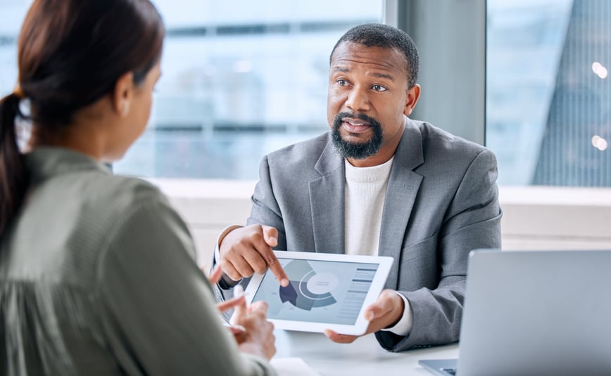 A businessman shows a data chart on a tablet to a colleague during a discussion in a bright office, indicating a professional meeting.-