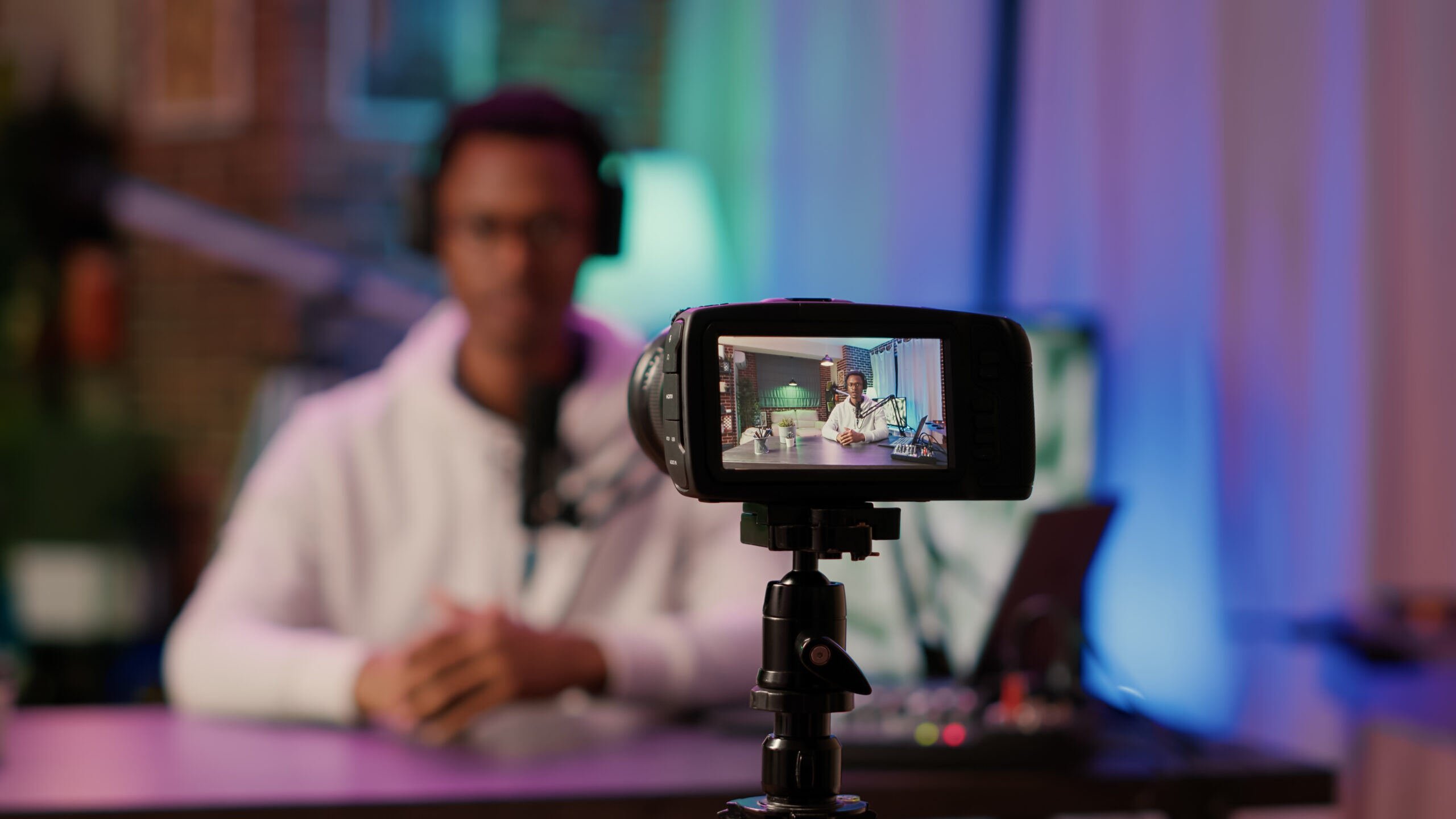A camera records a man with headphones at a desk with a microphone in a colorful, dimly lit studio. The camera screen shows him clearly.
