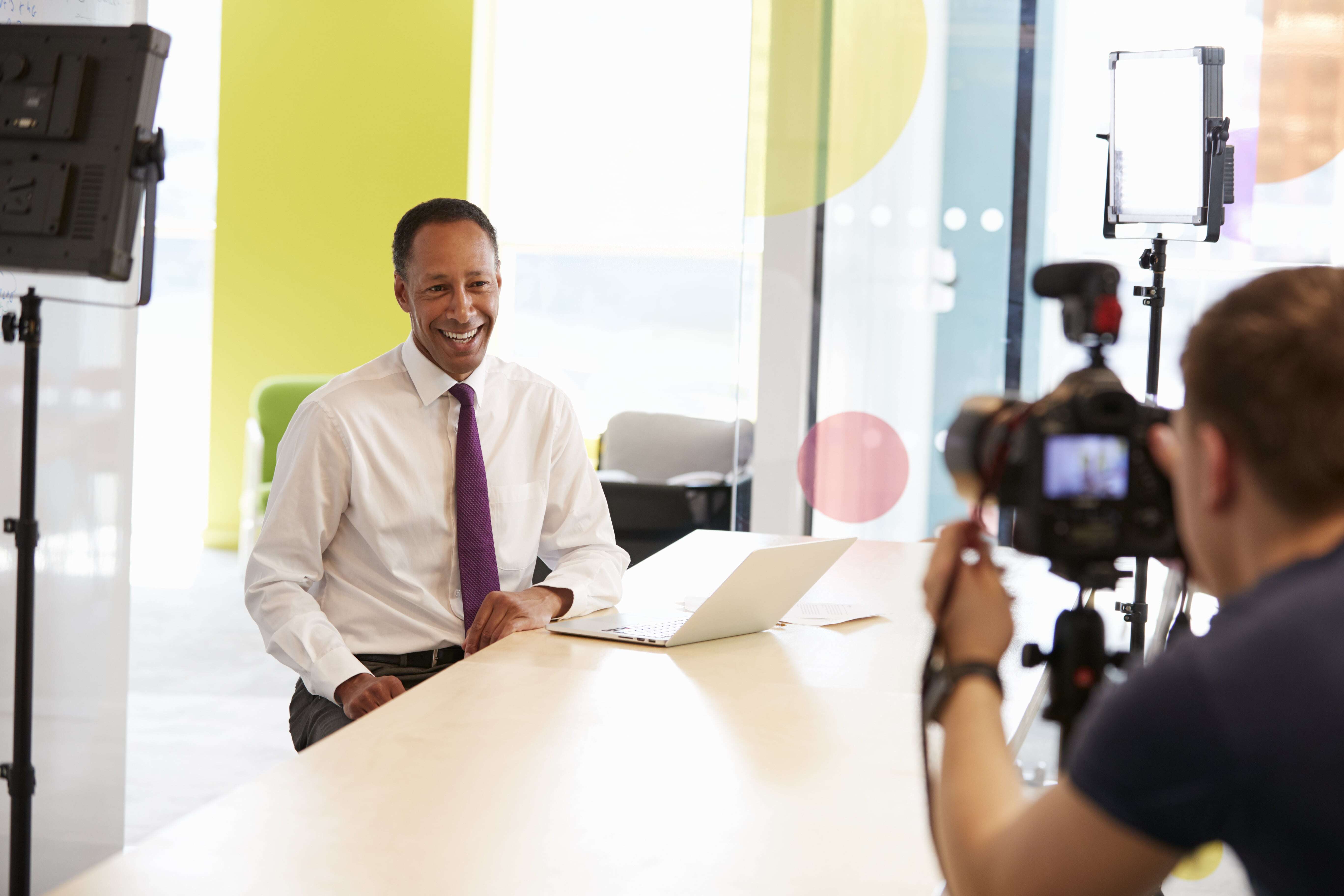 A cameraman recording a smiling businessman in a white shirt and purple tie during a corporate video shoot in a brightly lit office.