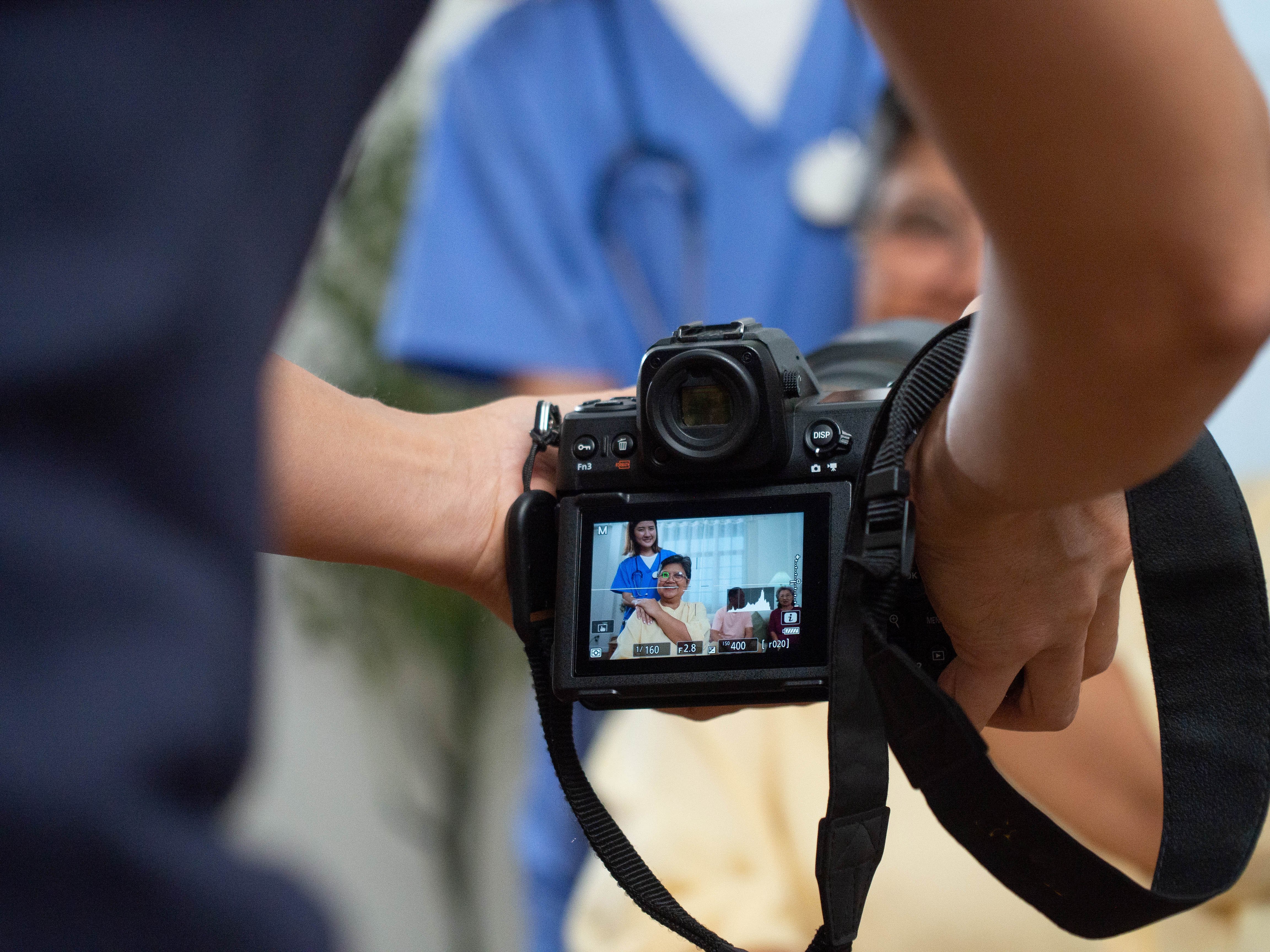 A close-up of a camera screen displaying a video being recorded featuring a group of people including a healthcare professional in scrubs and a patient