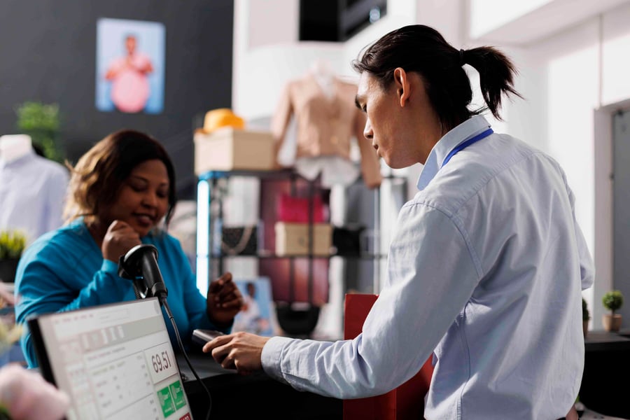 A customer in blue interacts with a cashier scanning an item, with a payment on the screen.