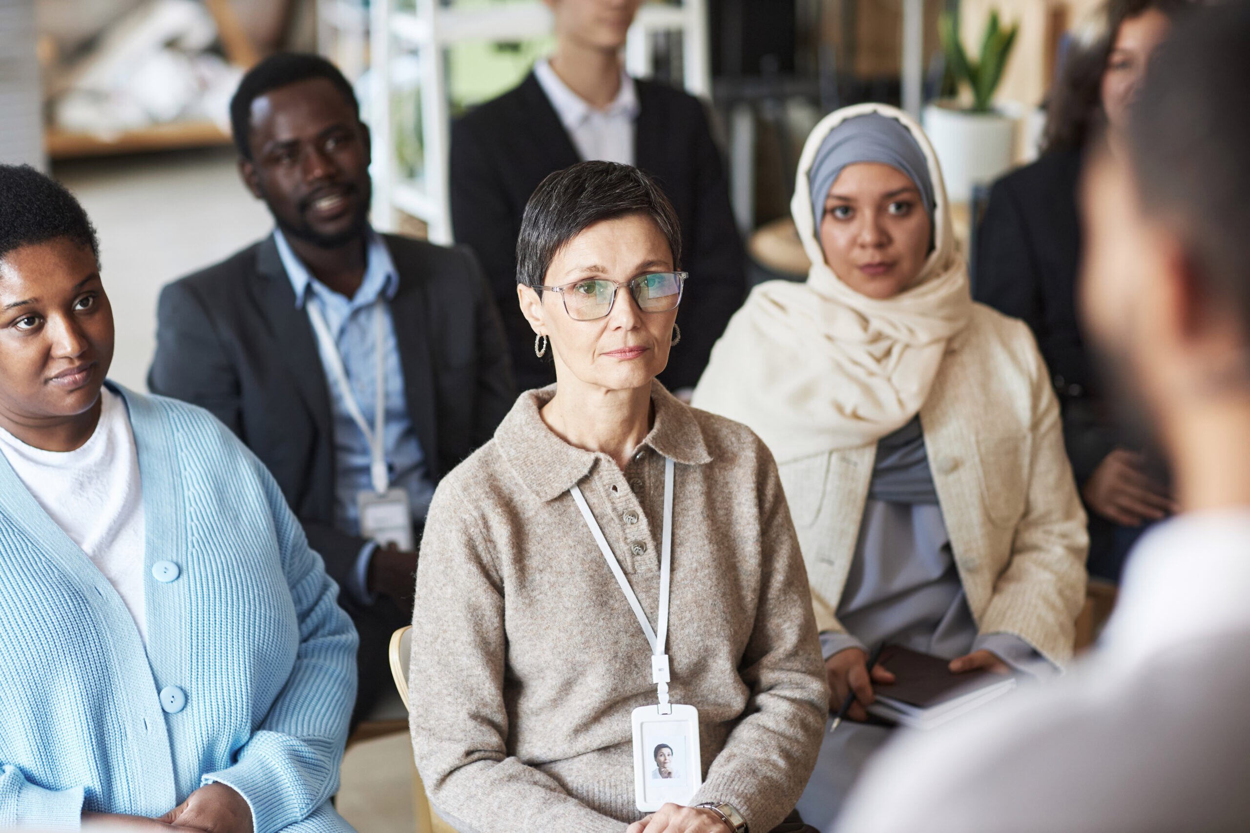A diverse group of marketers attentively listens to a speaker, with serious expressions and visible name badges, in a professional setting.