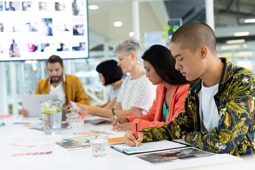 A diverse group of professionals sits at a table, reviewing documents and working on laptops in a bright, modern office setting.-