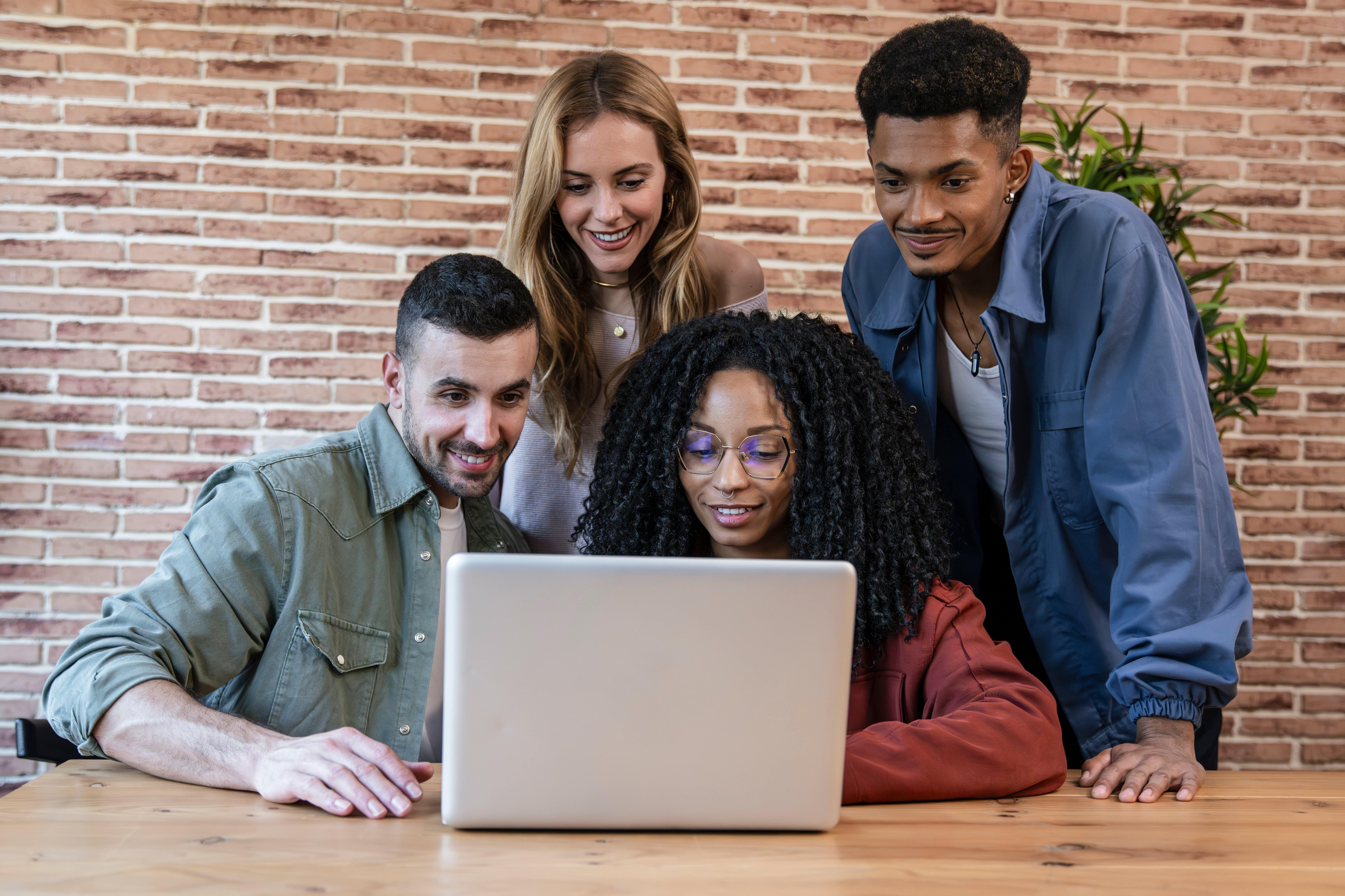 A diverse group of young adults huddled around a laptop, smiling and collaborating in a modern workspace