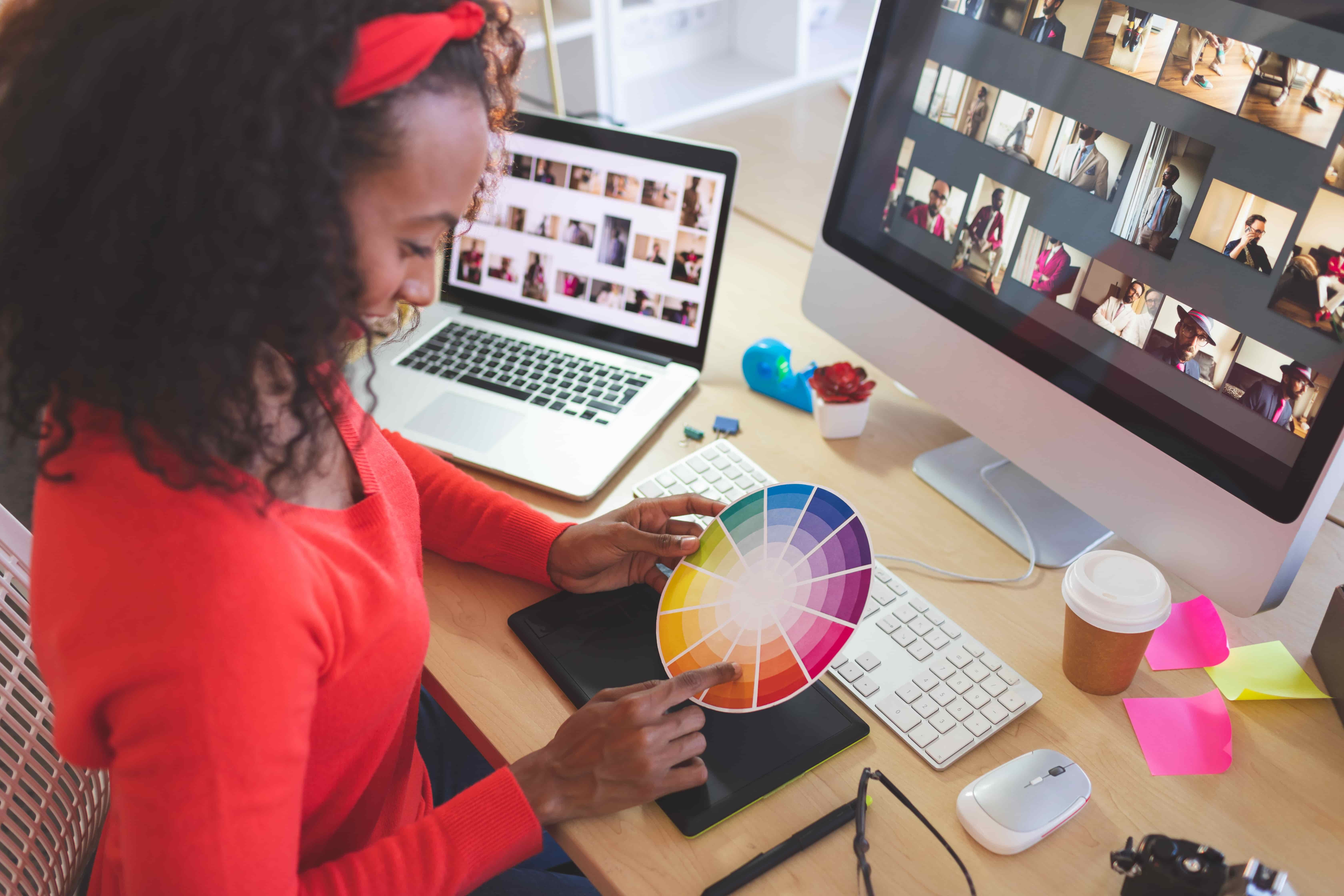 A graphic designer in a red sweater uses a color wheel at her desk, with multiple screens showing photo thumbnails and editing software.