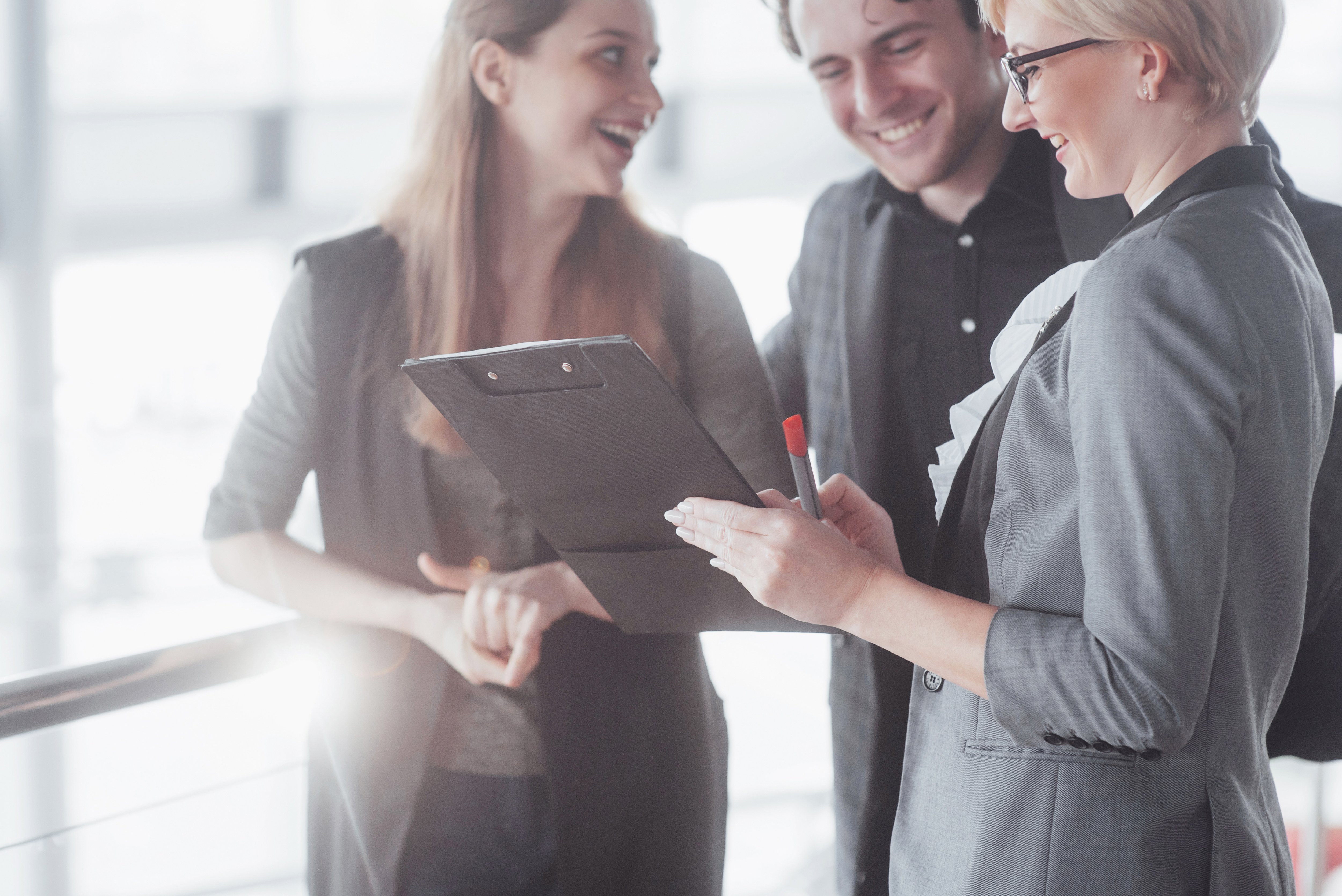 A group of business professionals smiling and discussing while one person holds a clipboard in a modern office setting.