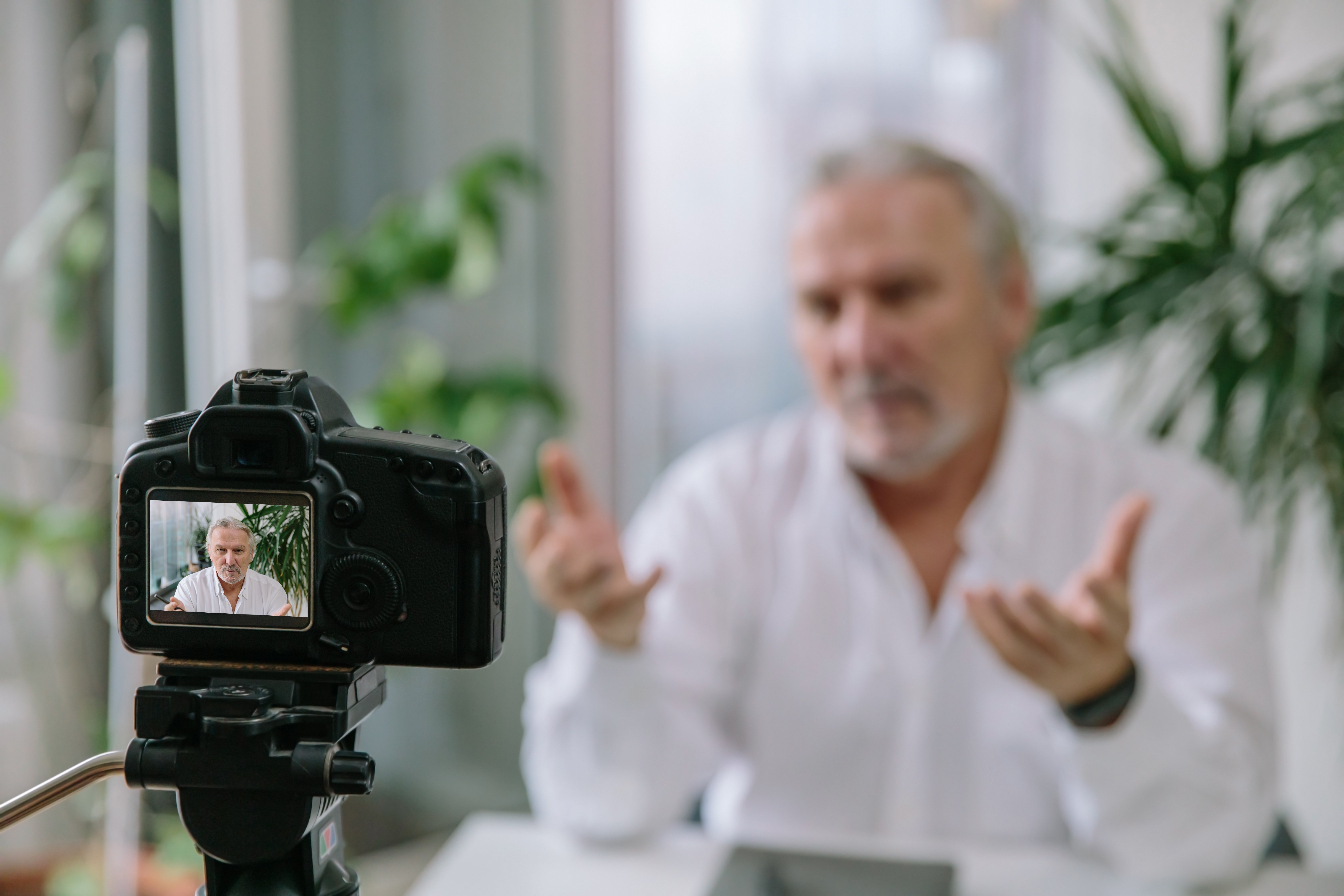 A man gestures while being recorded on a camera, with the camera screen showing him clearly in a bright room with plants in the background.