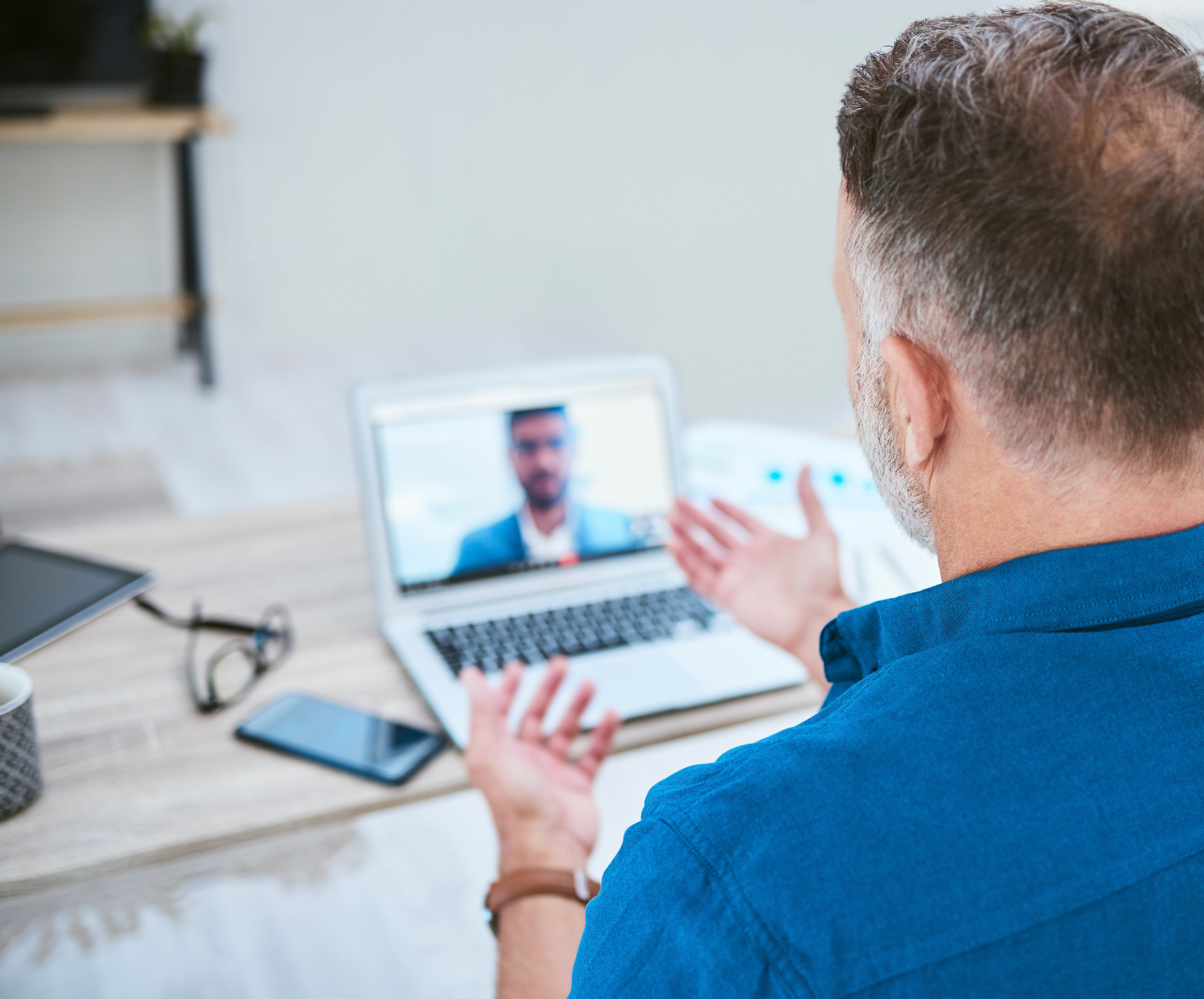 A man in a blue shirt gestures during a video call on a laptop, with a smartphone and glasses on the table nearby.