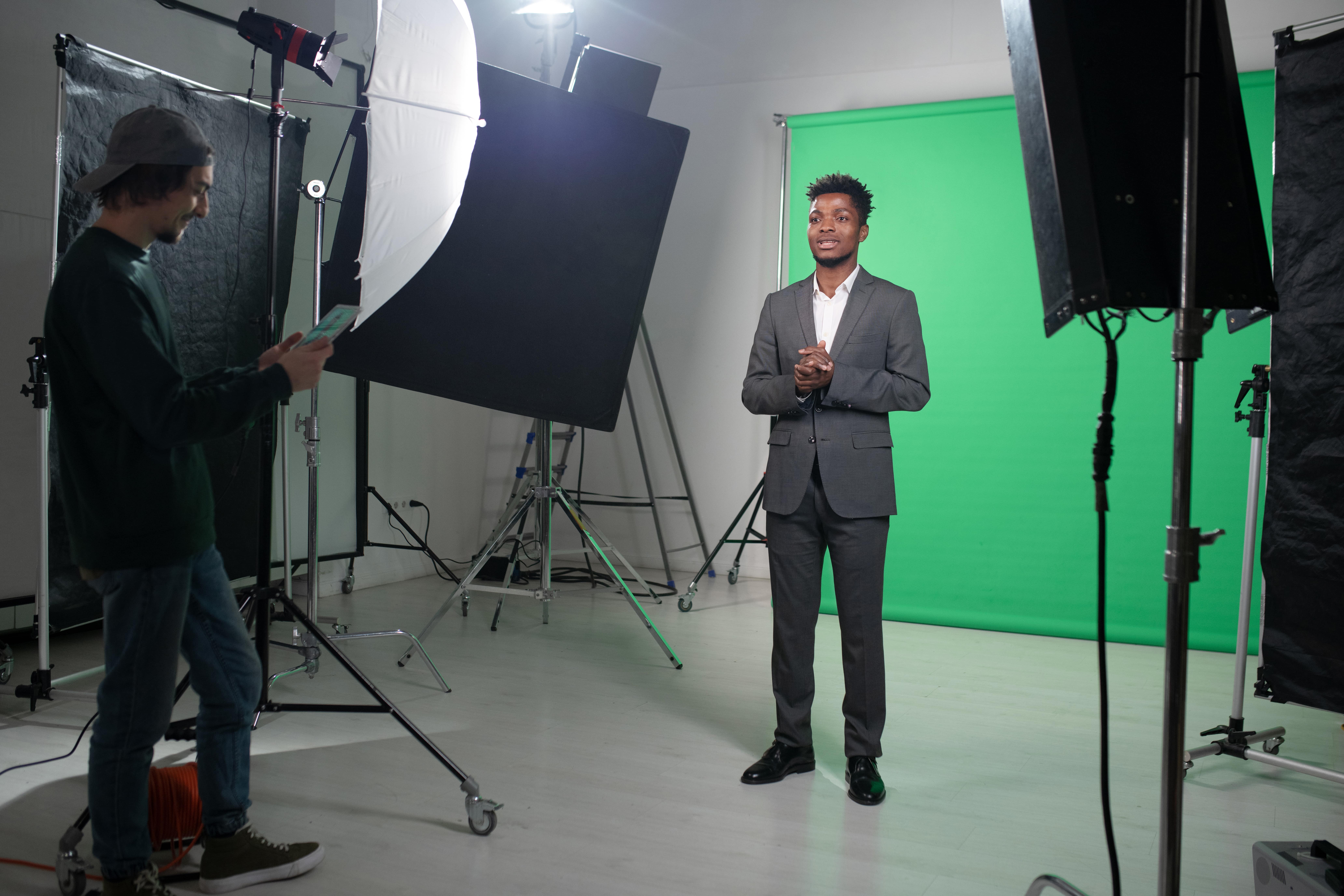 A man in a suit stands in a studio with a green screen backdrop, while a cameraman prepares to film