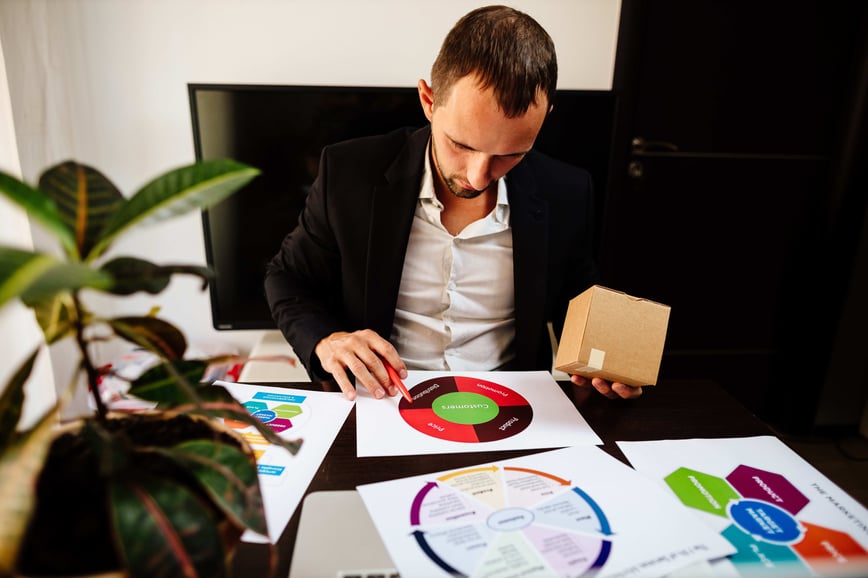 A man in a suit studies a circular marketing strategy diagram at a desk, holding a small cardboard box.-min