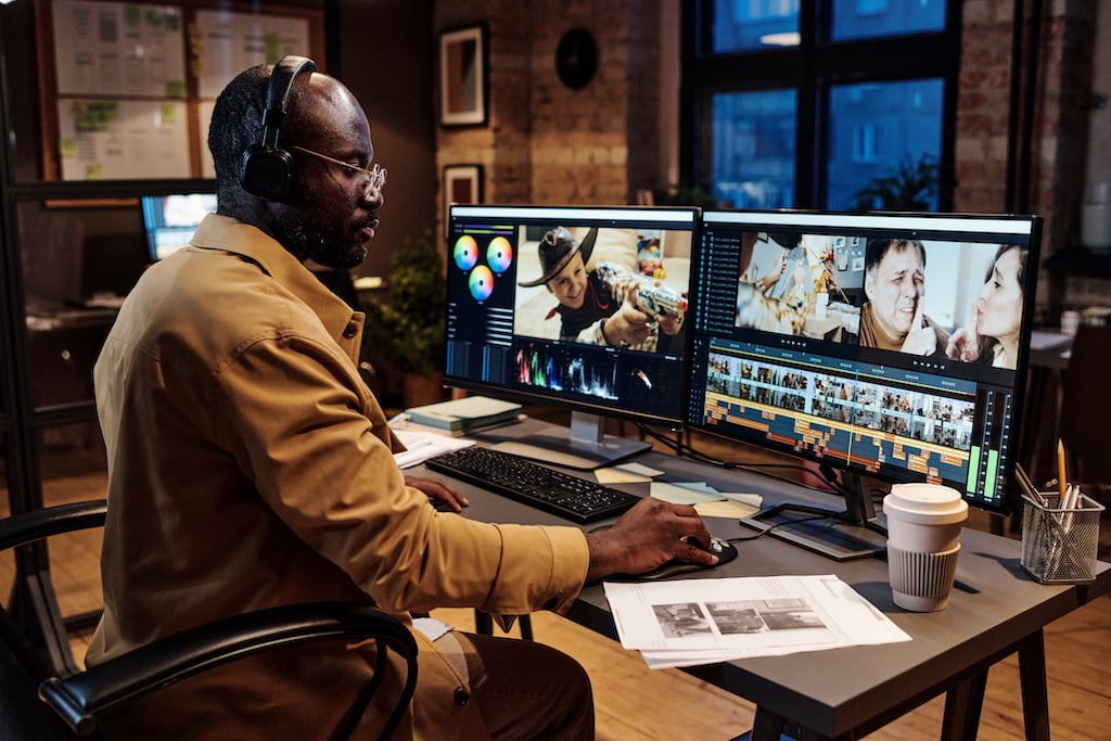 A man wearing headphones edits video on dual monitors in a dimly lit office, with coffee and documents on the desk