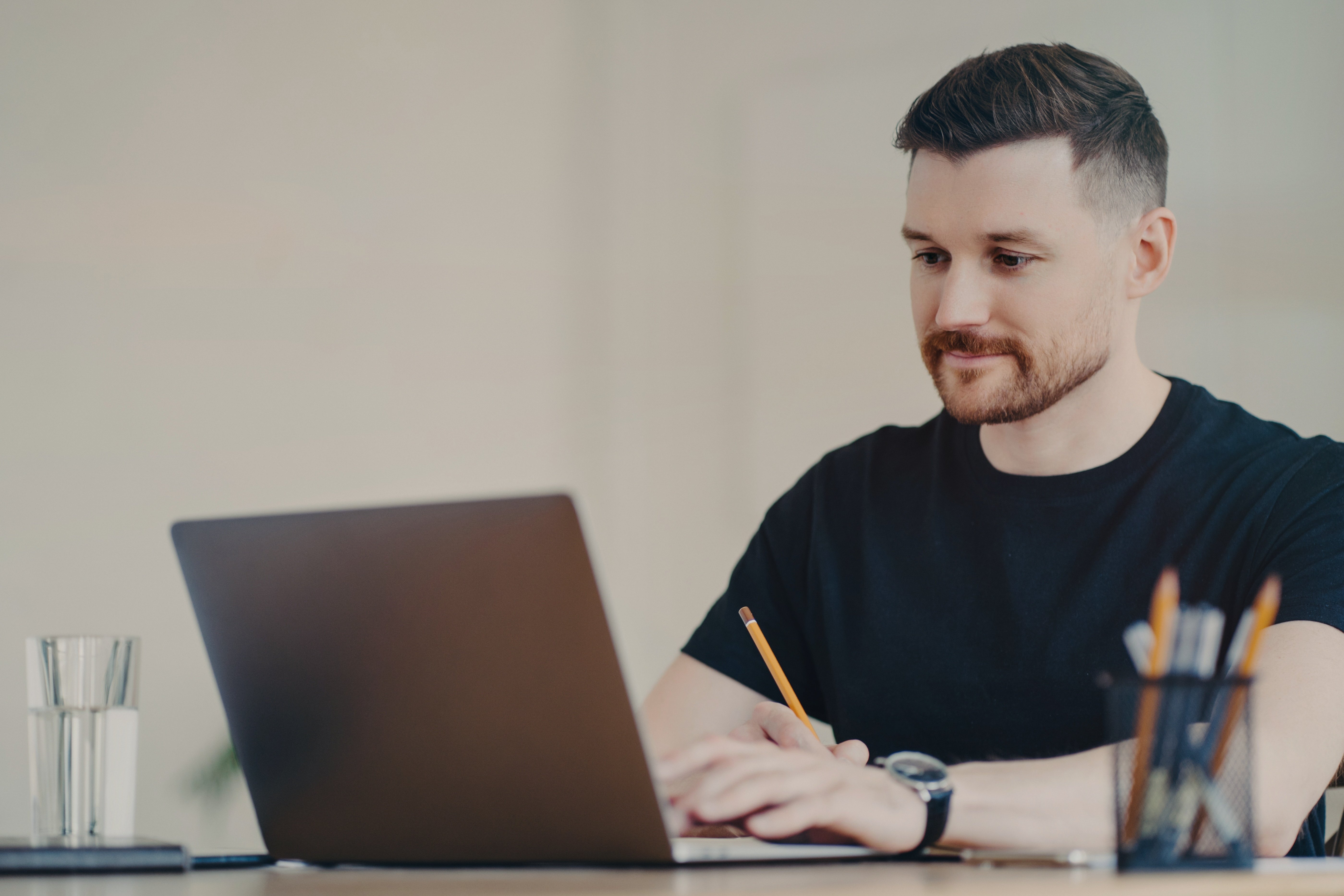 A man working on a laptop, holding a pencil, with a glass of water and a pen holder on the desk.