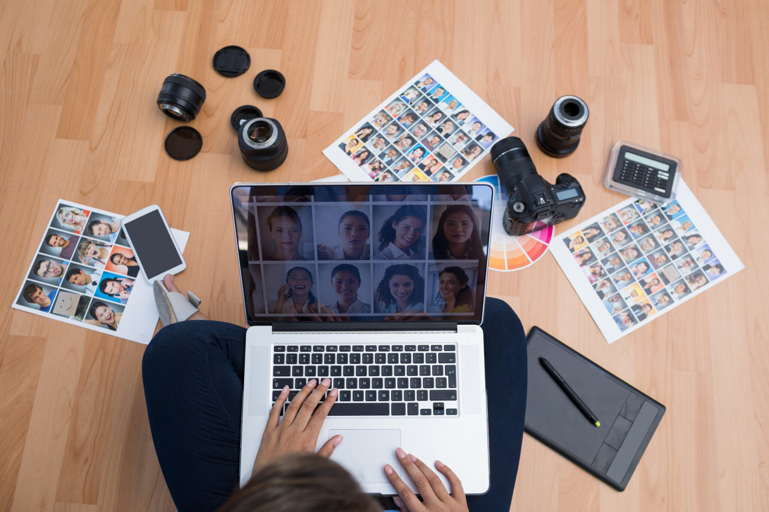 A person edits photos on a laptop, surrounded by camera lenses, a digital camera, and printed photo sheets on a wooden floor, indicating a photography or design project.