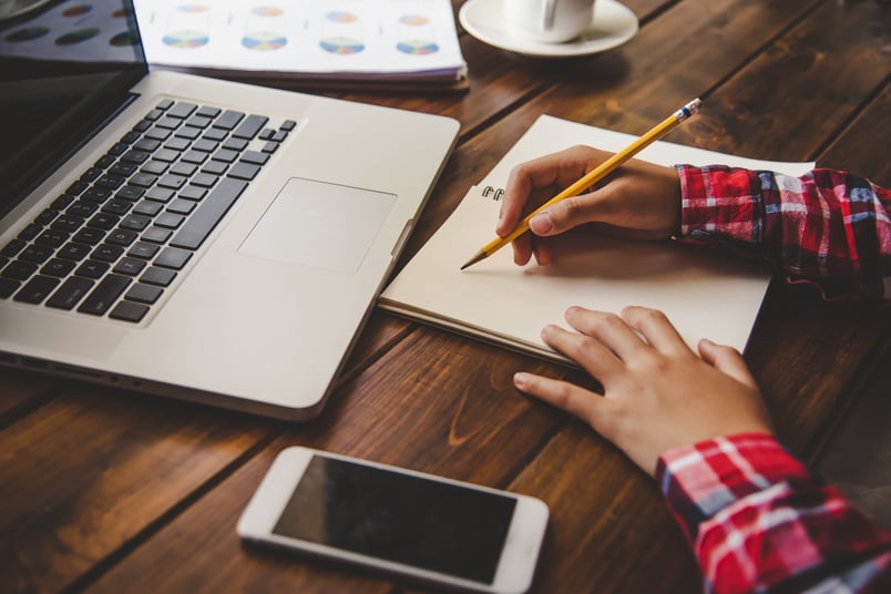 A person in a red plaid shirt writes in a notebook beside a laptop and smartphone on a wooden desk.-