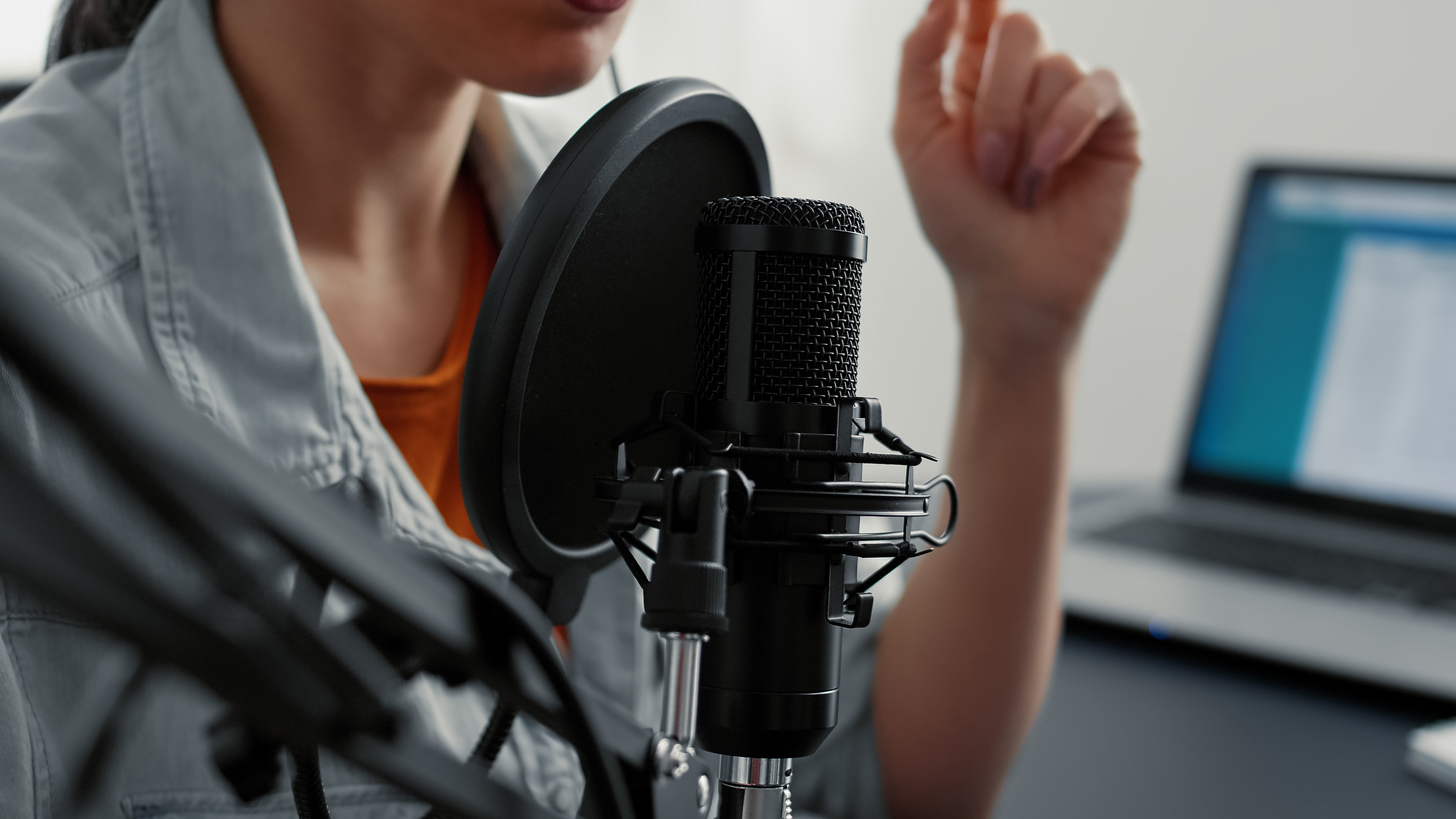 A person speaking into a microphone with a pop filter, possibly recording a podcast or live radio show, while working remotely with a laptop nearby.