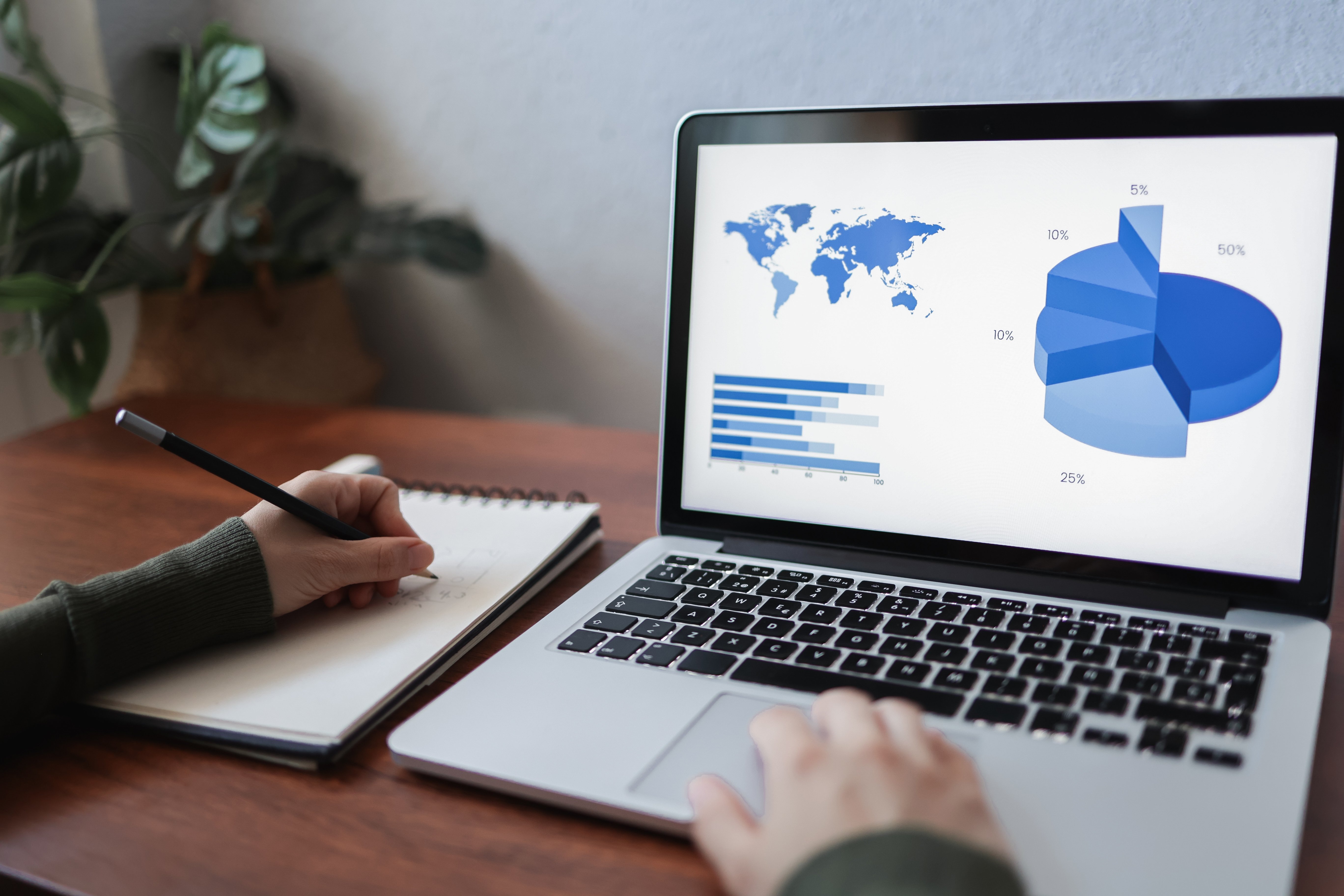 A person takes notes while analyzing data visualizations on a laptop, including a world map, bar chart, and pie chart.