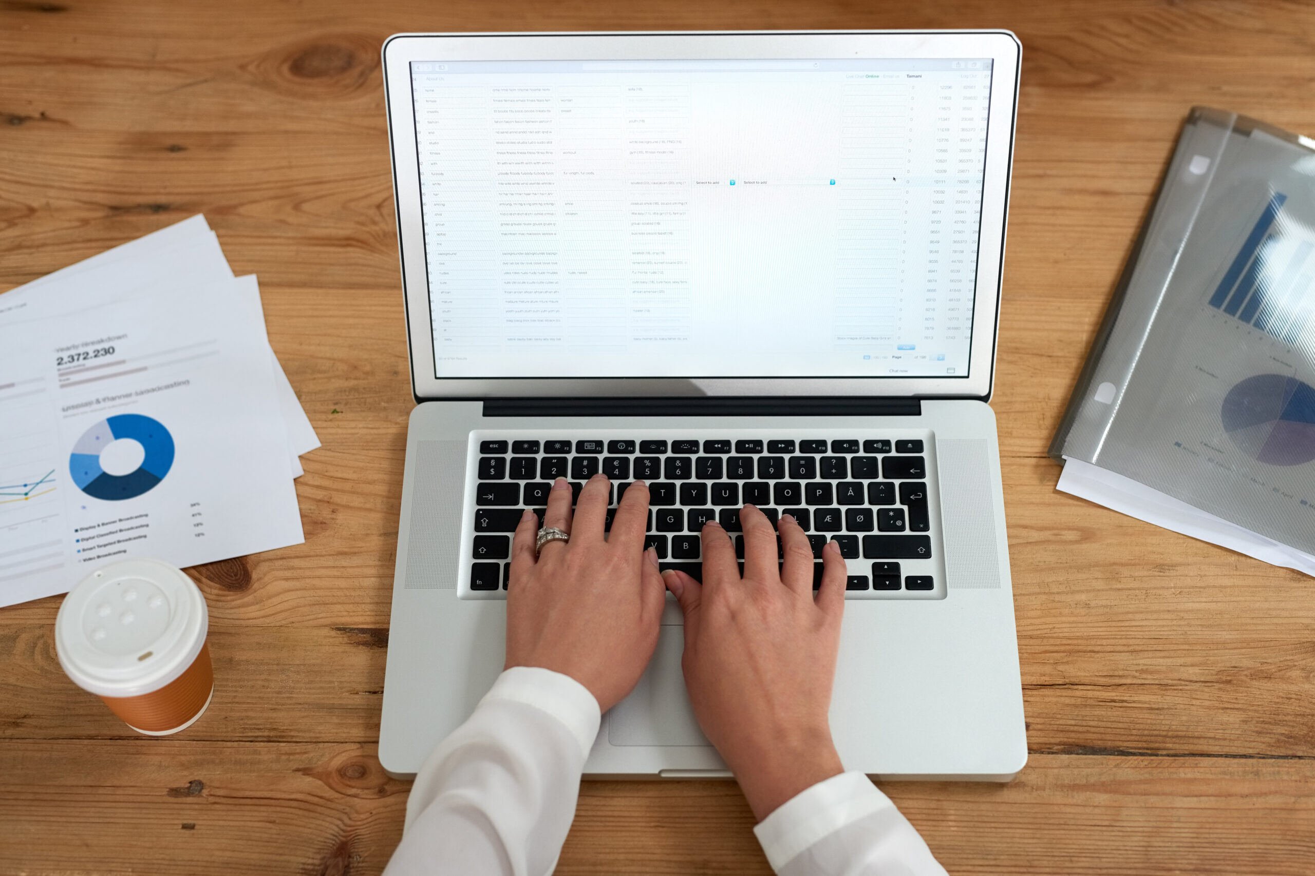 A person types on a laptop with analytical documents and a coffee cup on the wooden desk, focusing on work-related tasks with data sheets nearby.