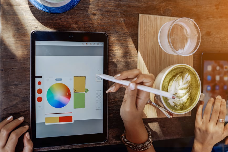 A person uses a stylus to design on a tablet with a colour wheel, while a cup of matcha latte and a glass of water sit on the table nearby.