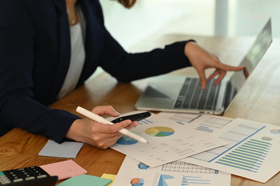 A person working at a desk with financial reports, using both a smartphone and a laptop to analyse data and make notes. 