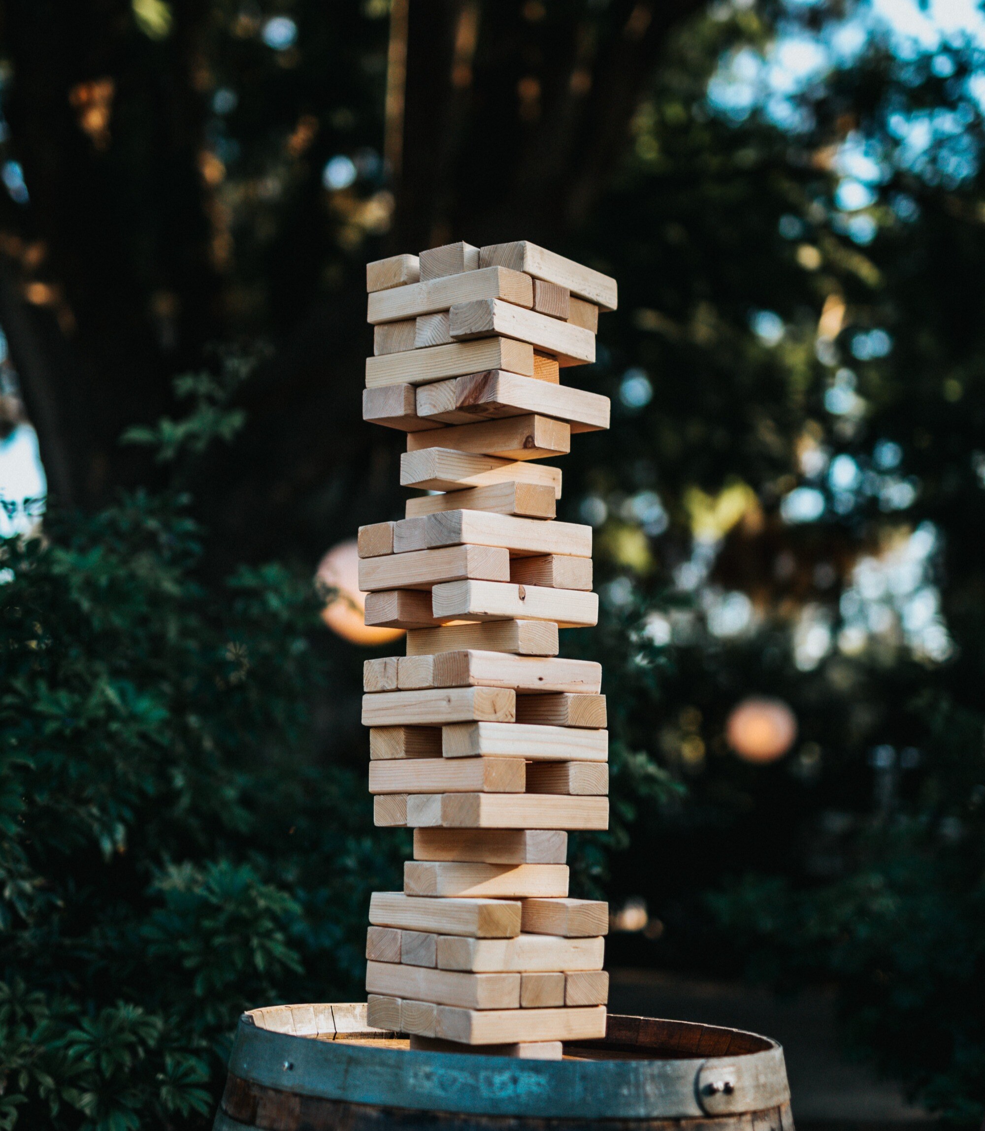 A tall, precariously stacked wooden block tower outside on a barrel, surrounded by greenery in a garden setting.