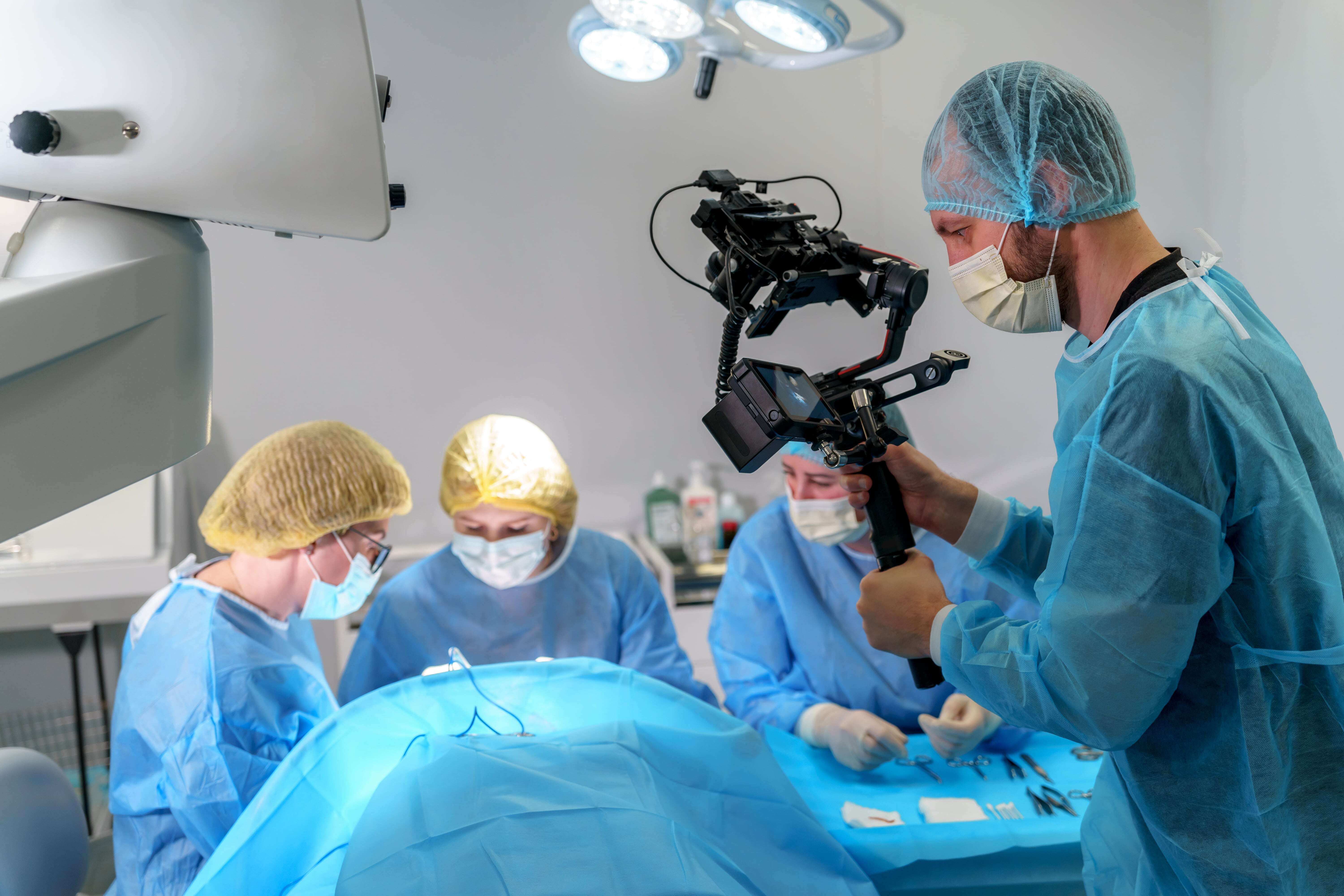 A videographer in surgical attire filming a team of surgeons performing an operation in an operating room.