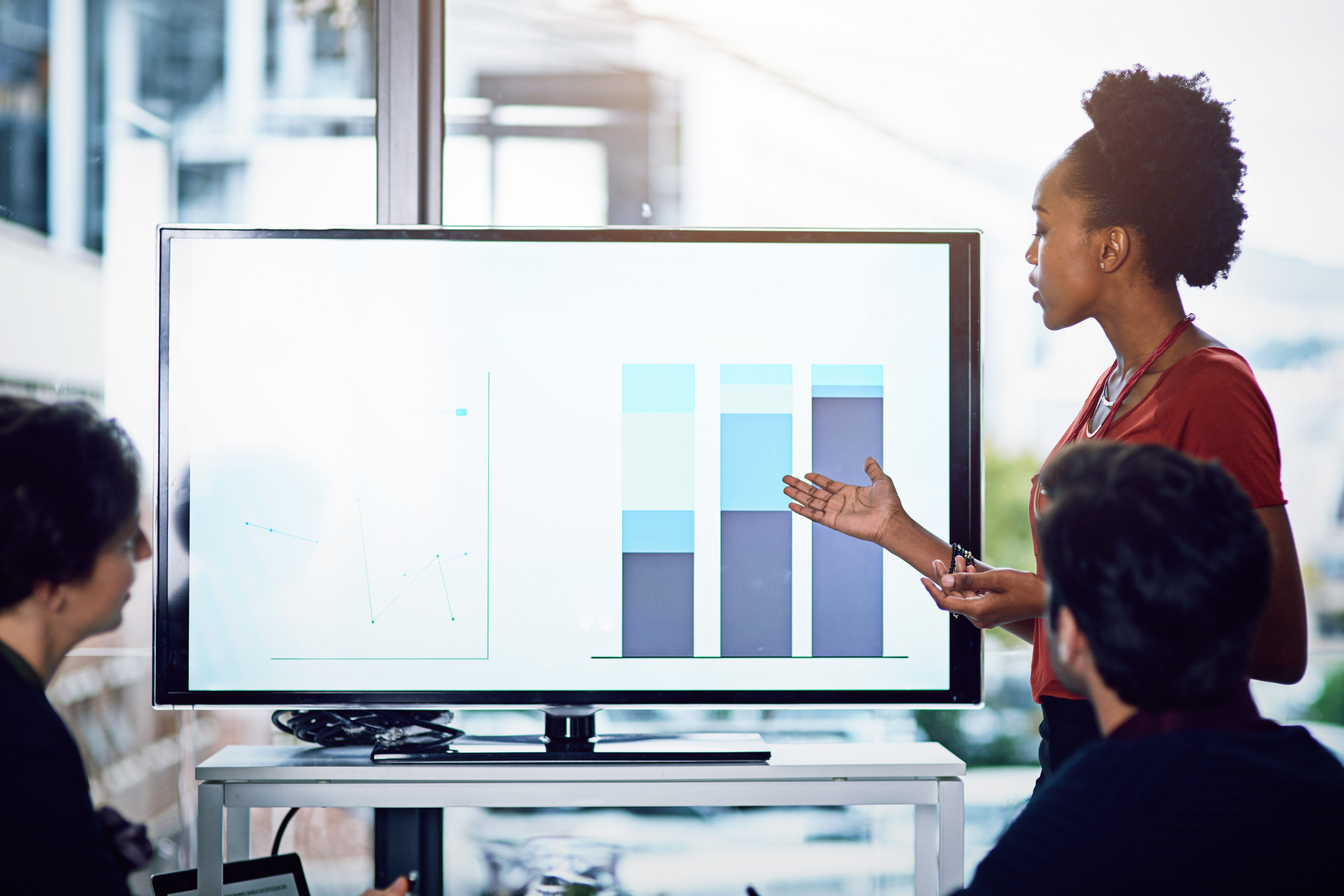 A woman gives a presentation to colleagues, pointing at bar graphs on a large screen in a modern office setting.