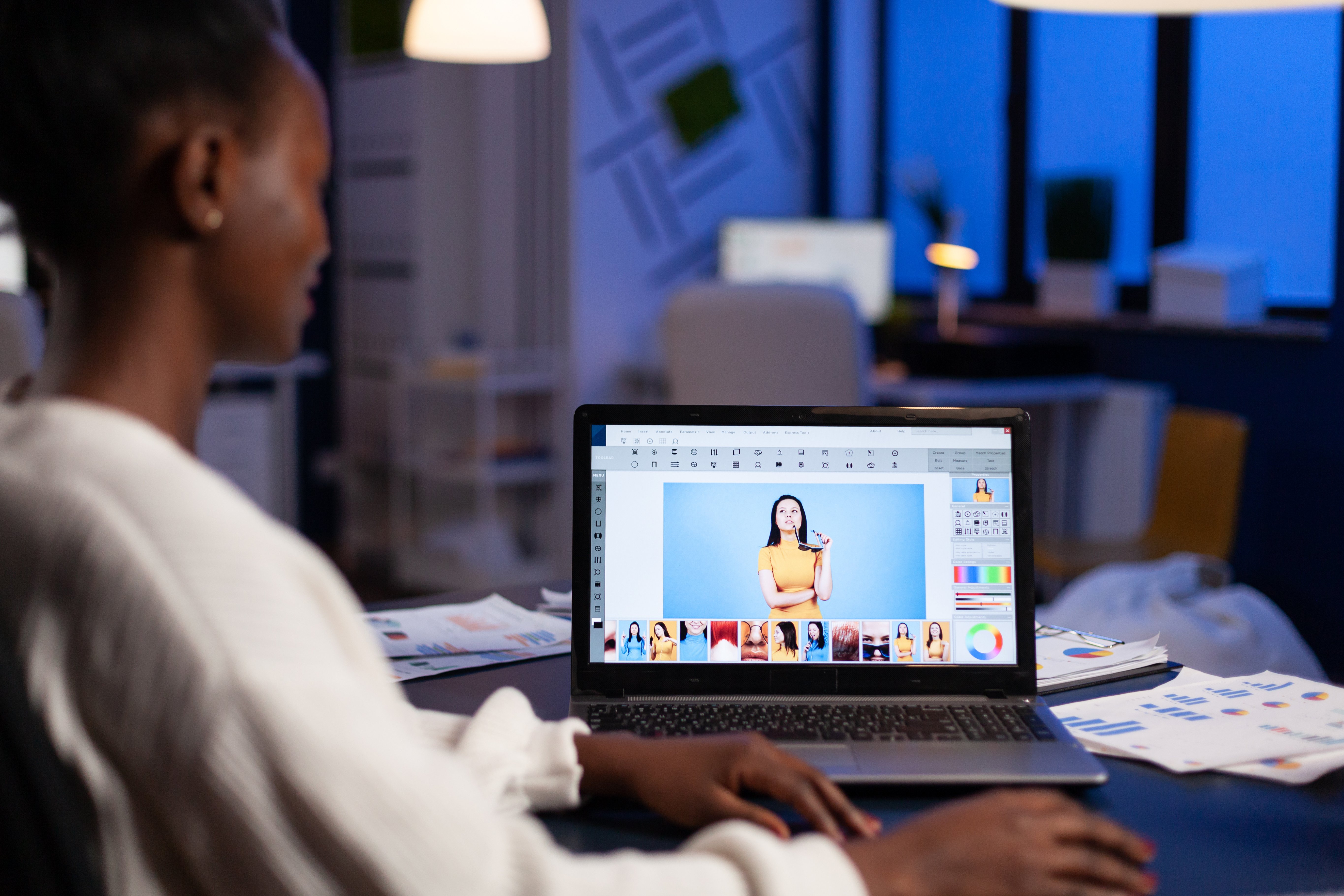 A woman working on a laptop in a dimly lit office, editing a photo of a woman in a yellow shirt using image editing software.-1