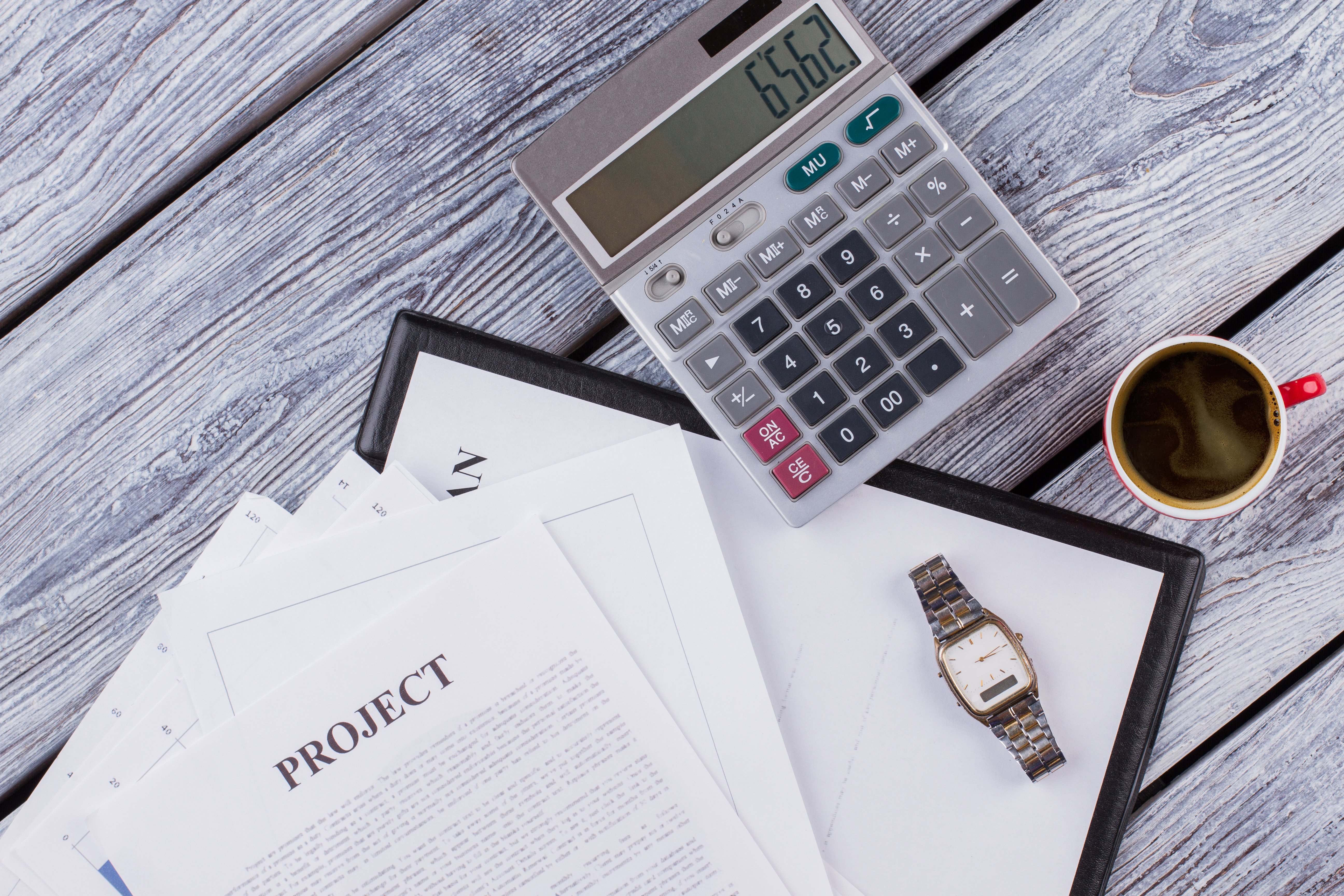 An office desk with a calculator, project documents, a watch, and a cup of coffee on a wooden surface.