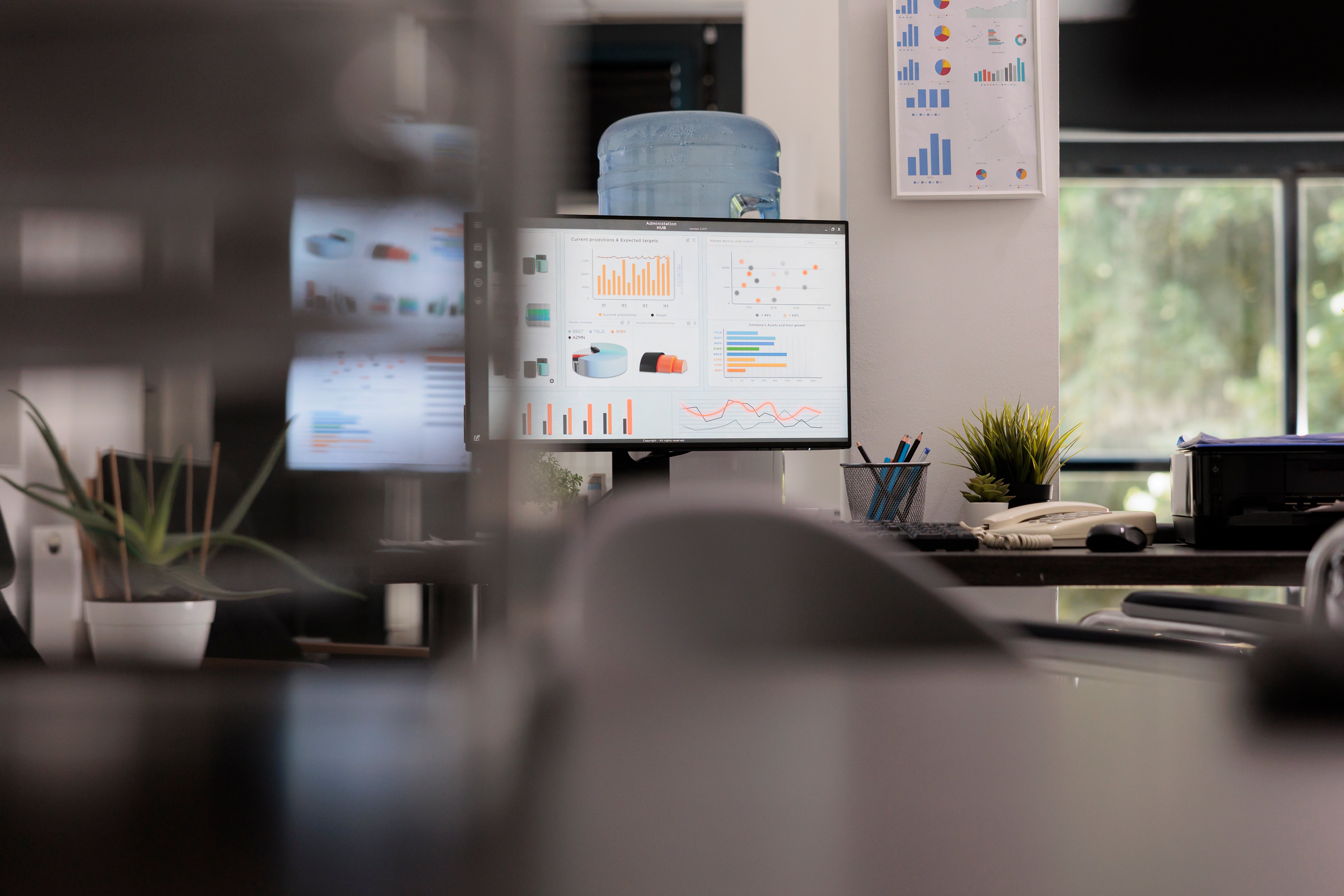 An office desk with dual monitors displaying charts and graphs, with a water cooler and plants in the background, suggesting a work setting.