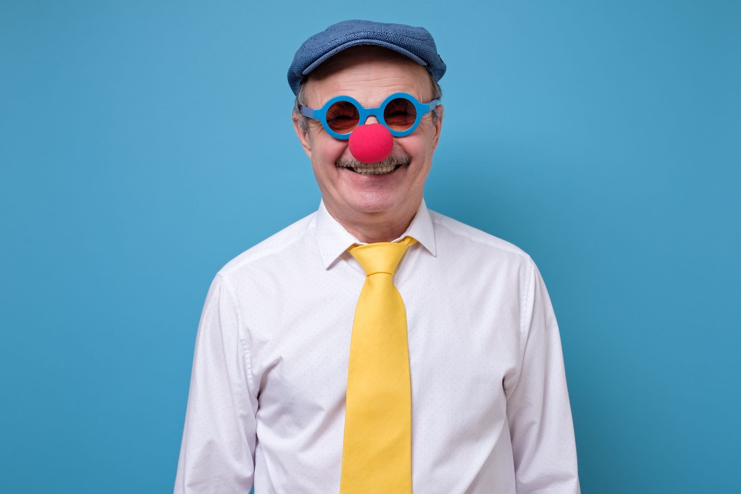 Bearded man in a yellow tie wearing a clown nose and blue glasses, smiling.-min