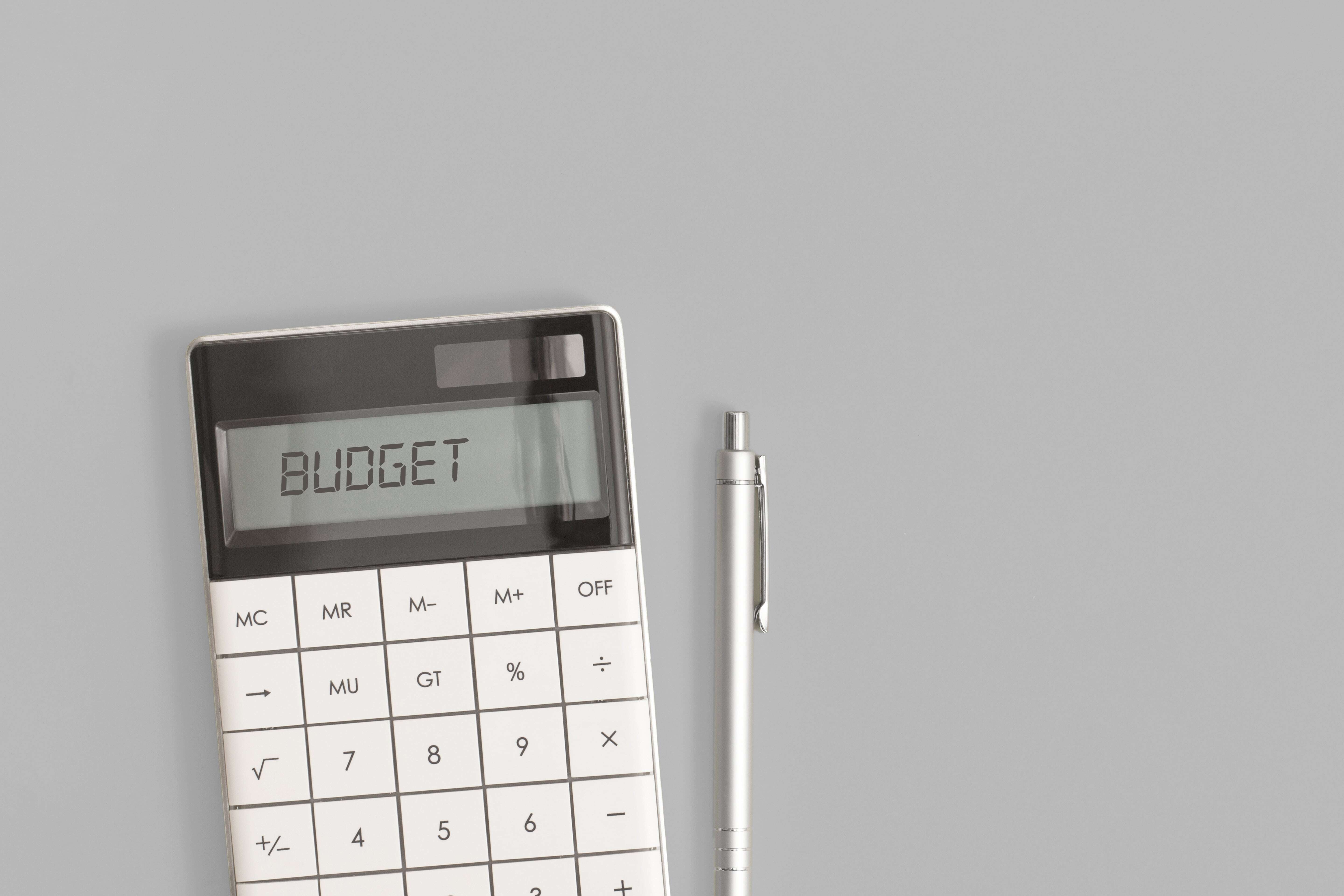 Calculator displaying the word BUDGET on a grey background with a silver pen placed next to it.