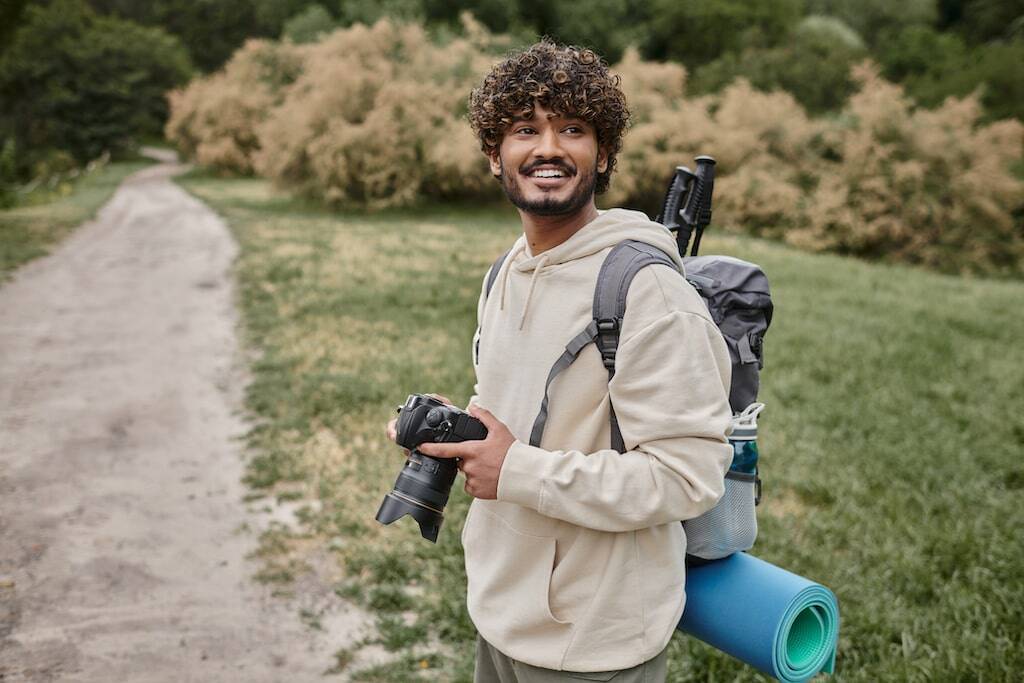 Cheerful man with curly hair holding a camera, wearing a backpack and hiking gear on a nature trail.-min