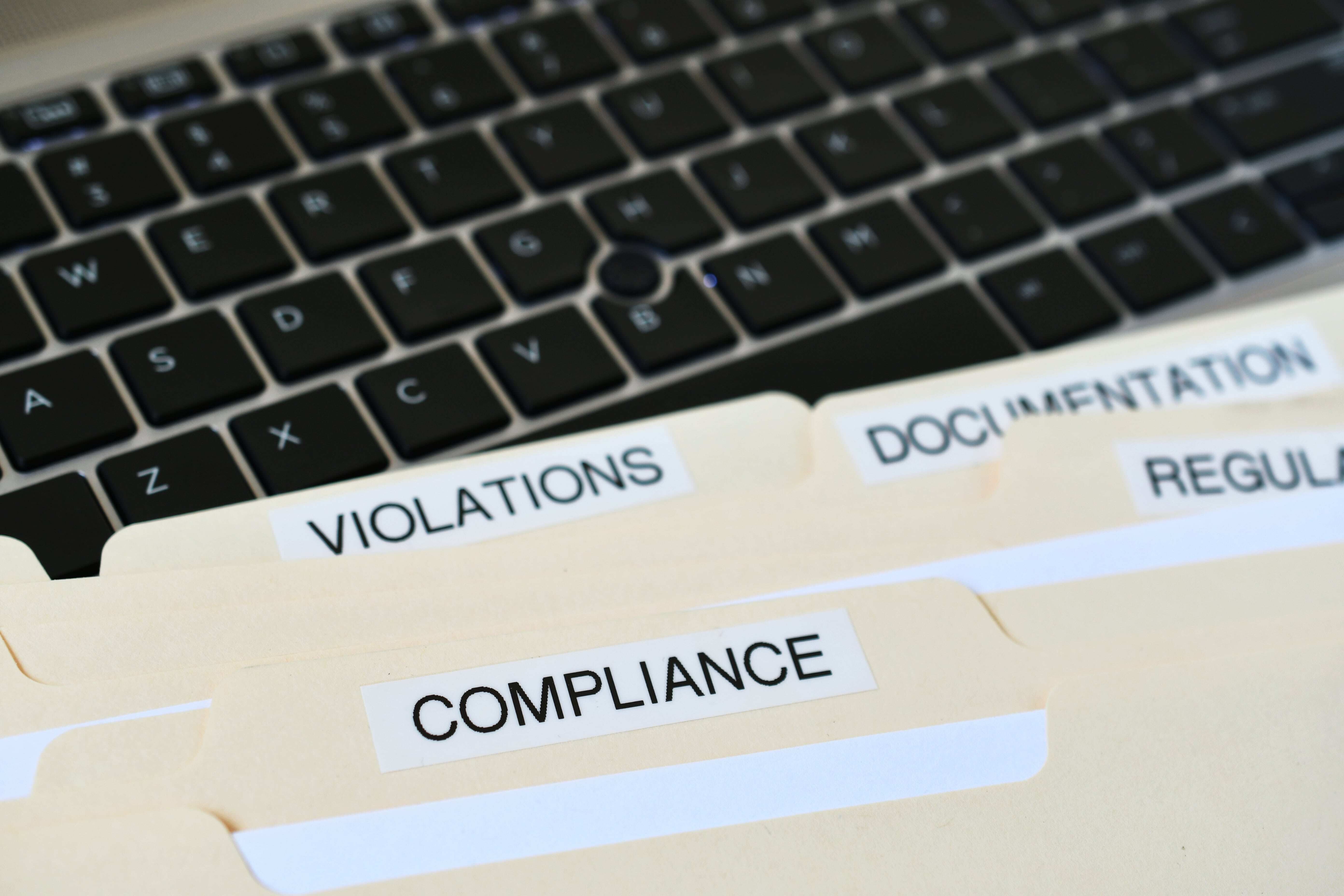 Close-up of a laptop keyboard with four manila folders labeled Violations, Compliance, Documentation, and Regulations placed on top.-min