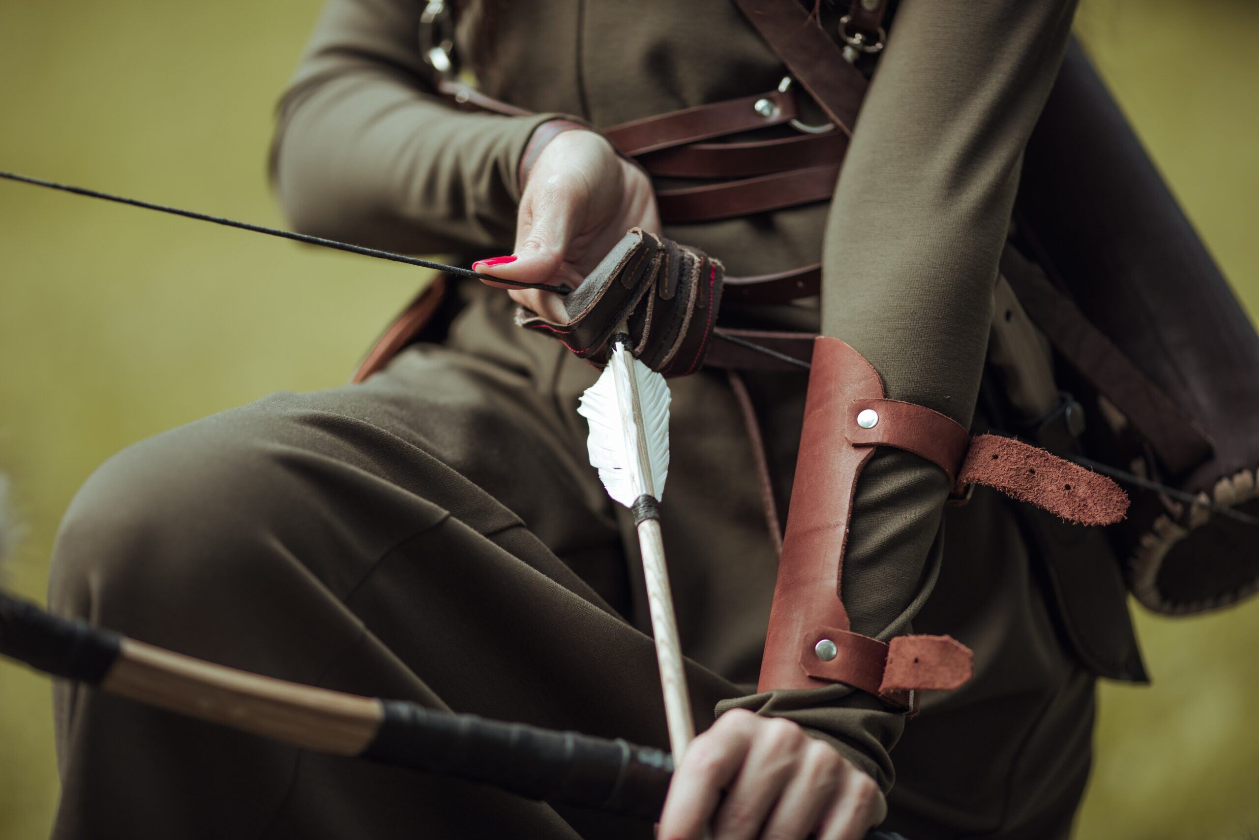 Close-up of a woman drawing a bowstring with an arrow, wearing green and leather armguards.-min