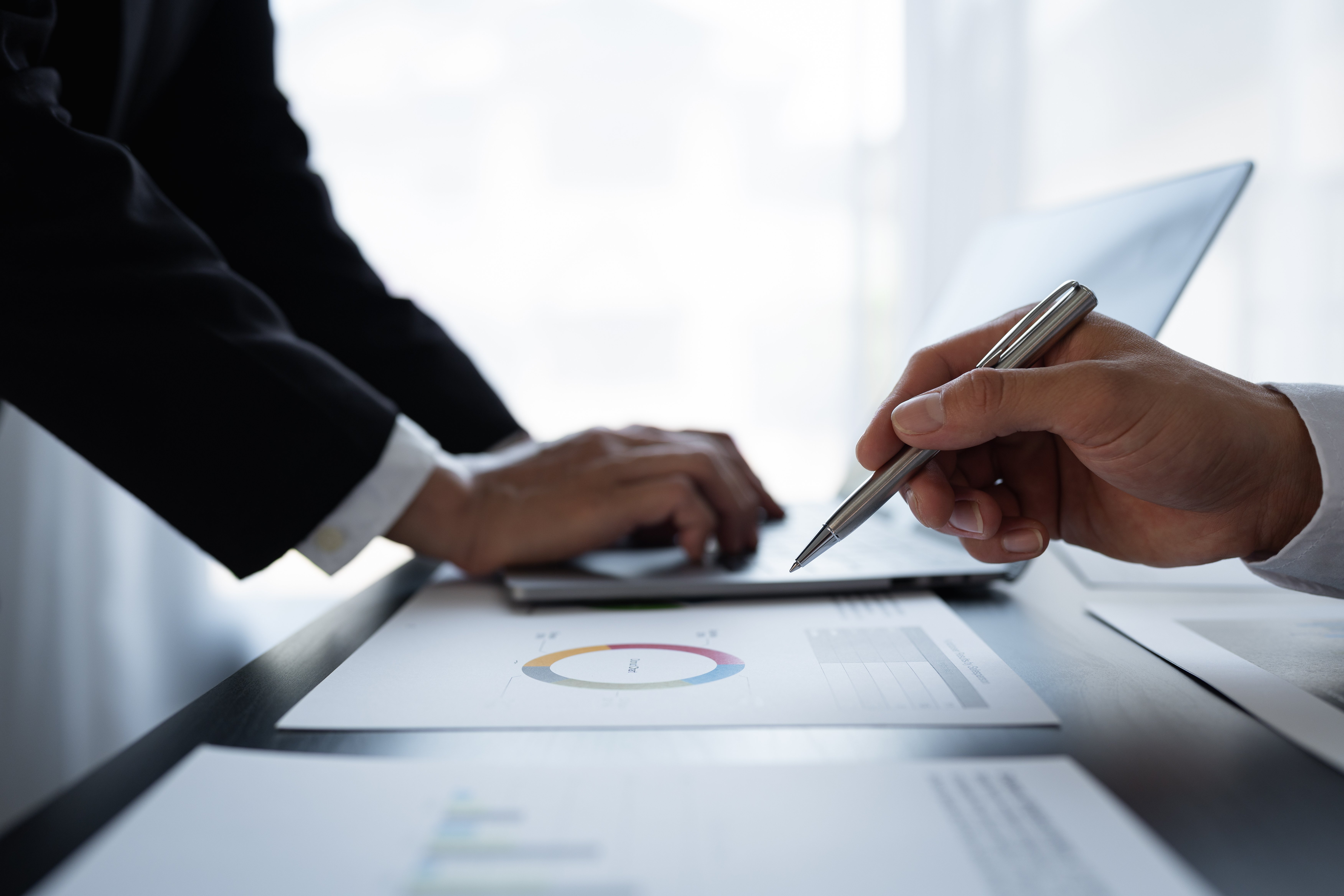 Close-up of two business professionals analysing charts with a laptop in the background, one holding a pen pointed at a pie chart.