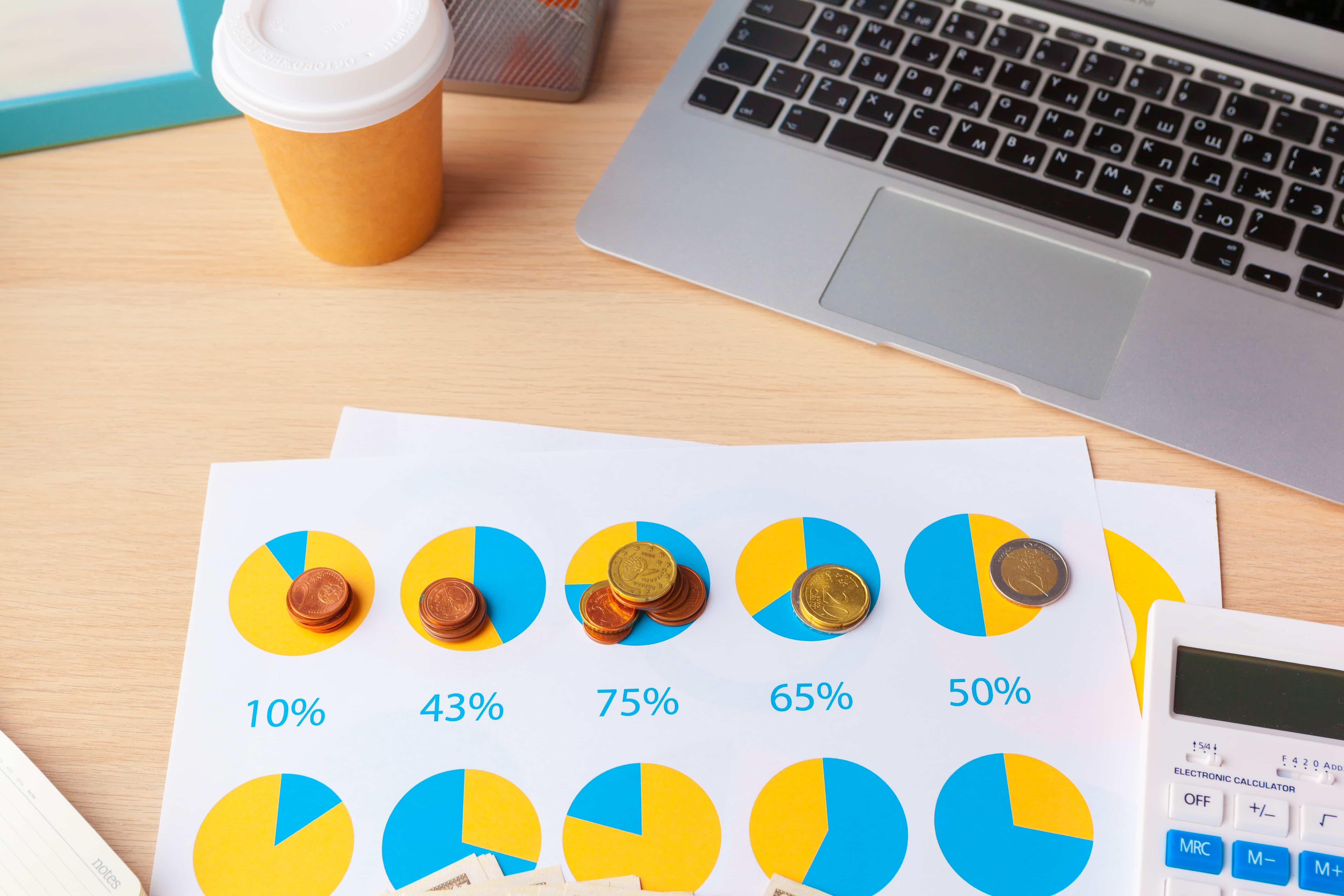 Coins on pie charts showing percentages, next to a laptop and calculator on a desk, suggesting financial analysis or budgeting.