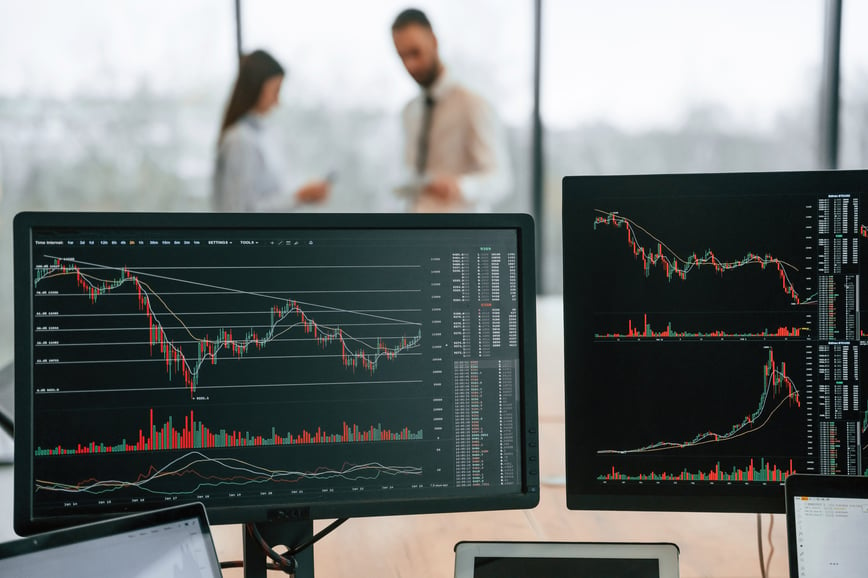 Computer monitors display complex data charts and trends related to the medical device industry, while two professionals discuss strategy in the background-