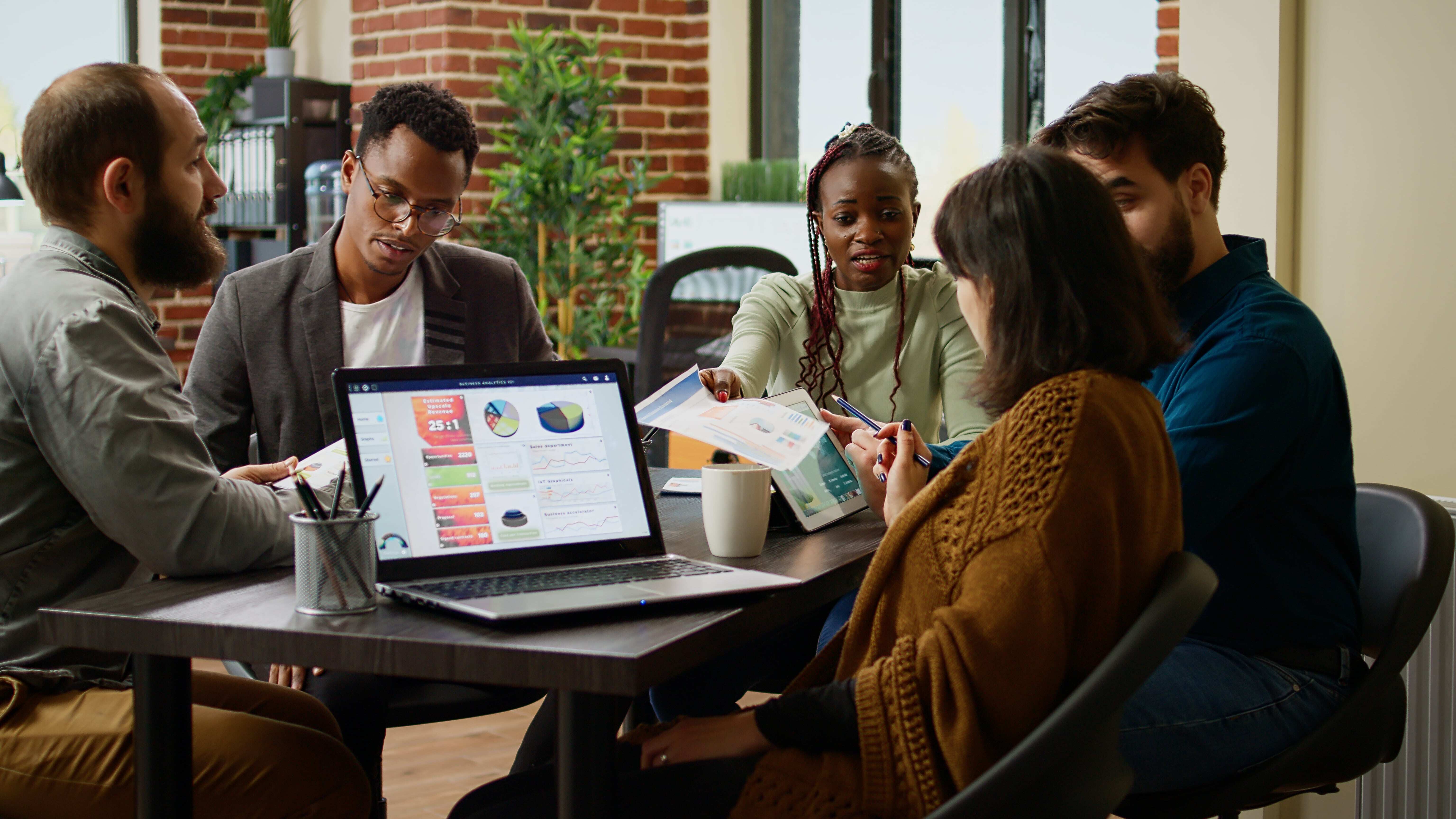 Diverse team of colleagues in a meeting, discussing documents and data displayed on a laptop in a modern office