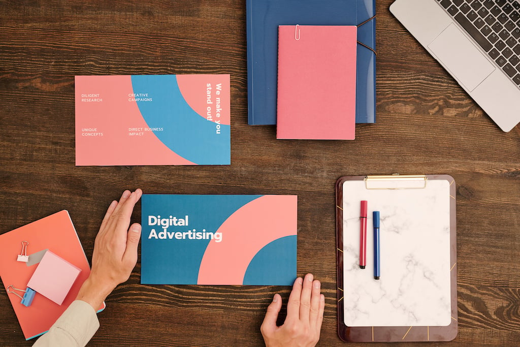 Hands organise a Digital Advertising brochure on a desk with notebooks, a clipboard, and a laptop, emphasising creative campaigns