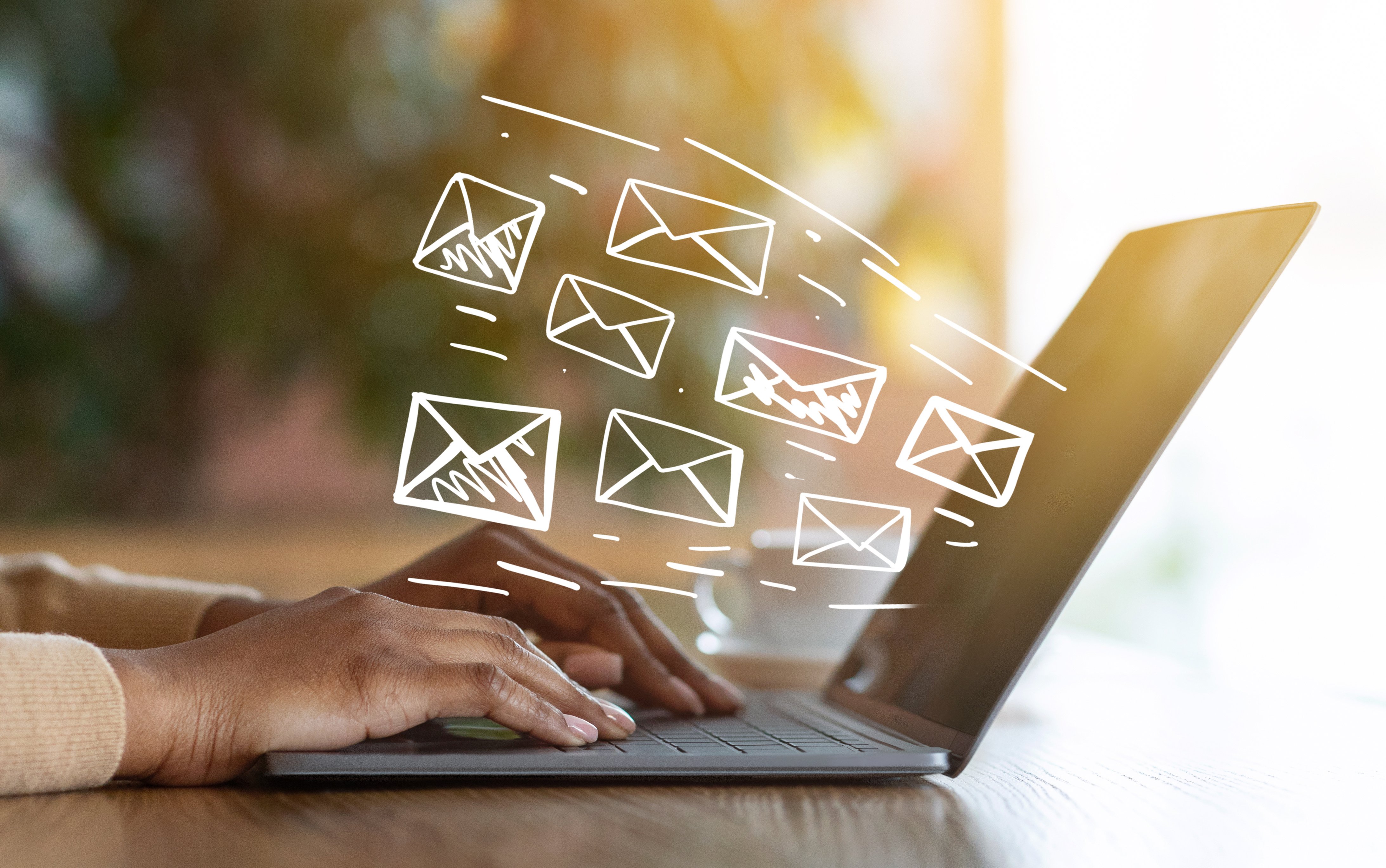 Hands typing on a laptop with white envelope icons floating above, representing emails being sent. Sunlight softly illuminates the scene.