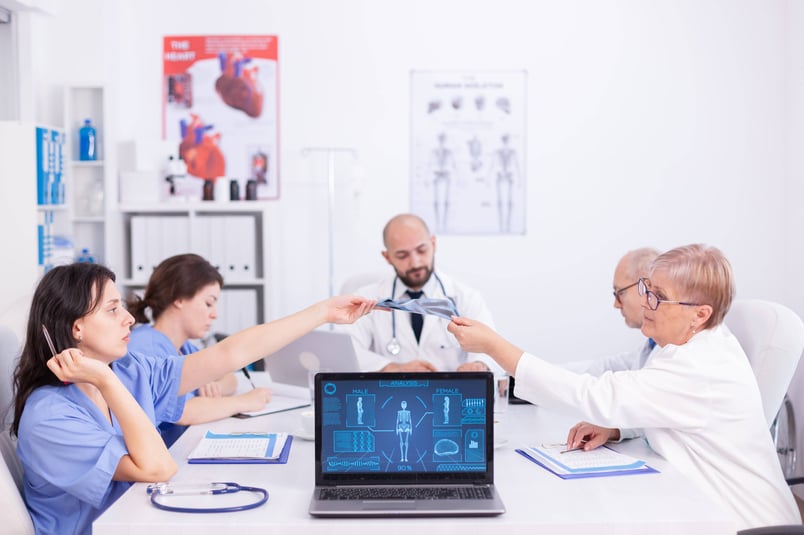 Healthcare professionals sat in doctors office having meeting, with a medical device website on a laptop screen