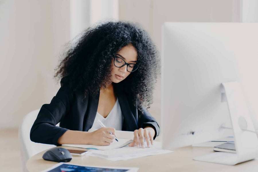 Woman sits in front of computer monitoring, writing an article in a notebook, which she will optimise to rank highly on search engines. 