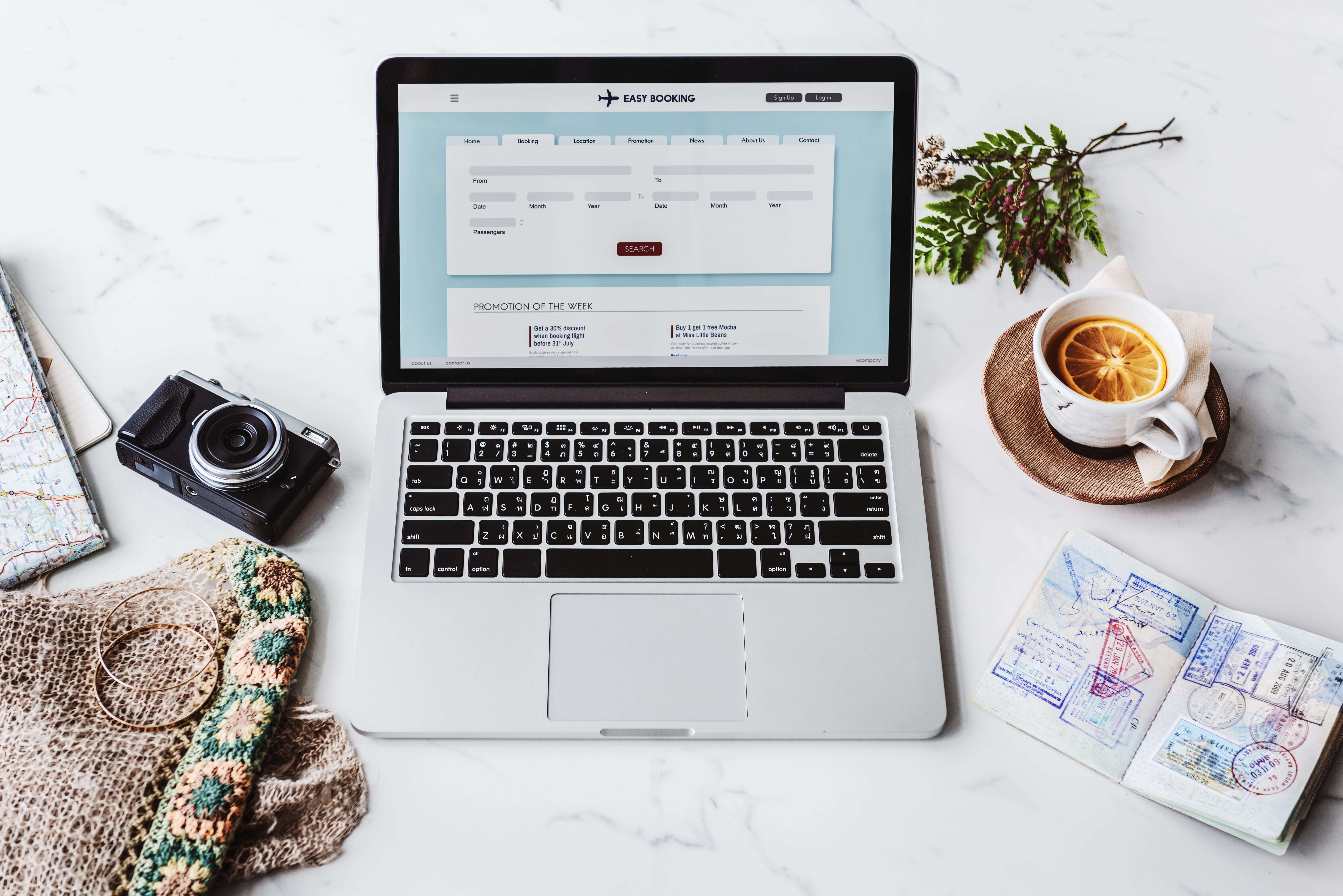 Laptop on a marble surface displaying an online booking website, surrounded by a camera, map, passport with stamps, and a cup of tea with an orange slice