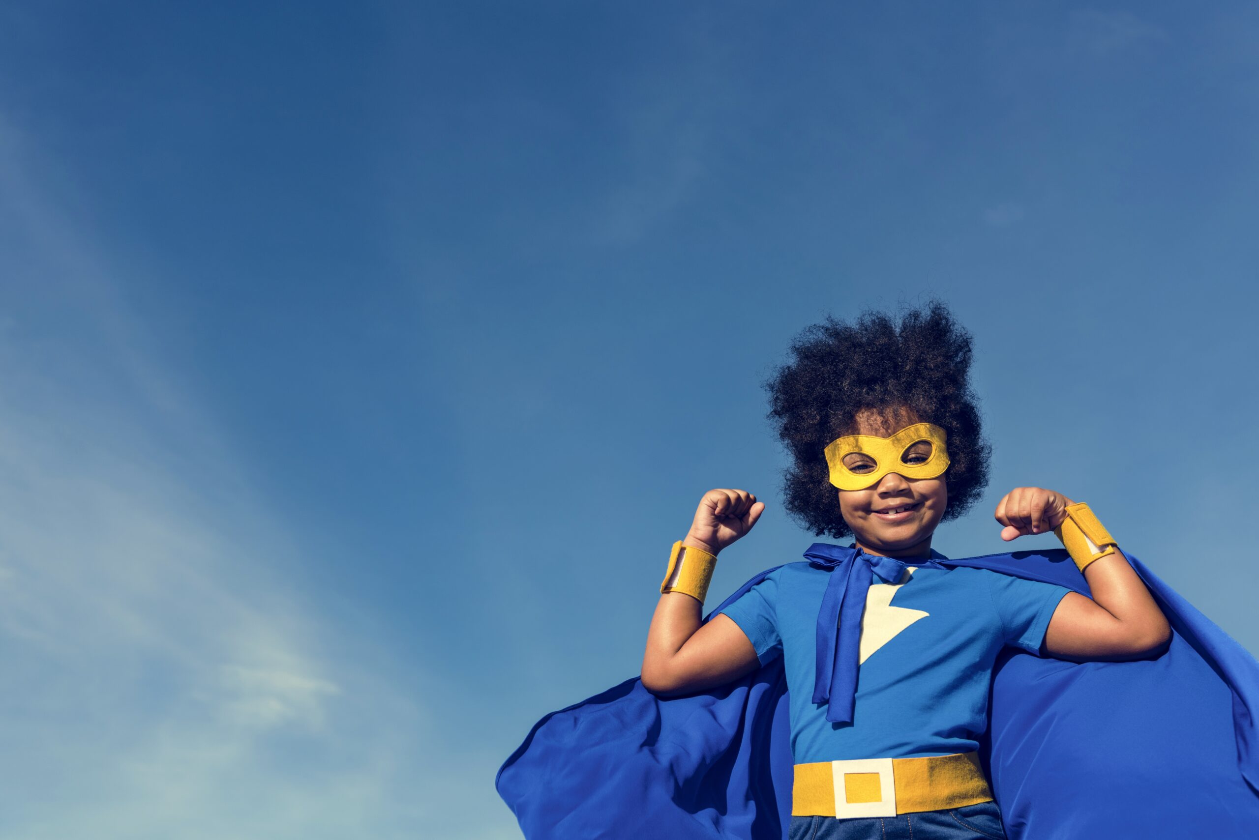 Little boy dressed as a superhero in blue, flexing his muscles against a clear blue sky.-min