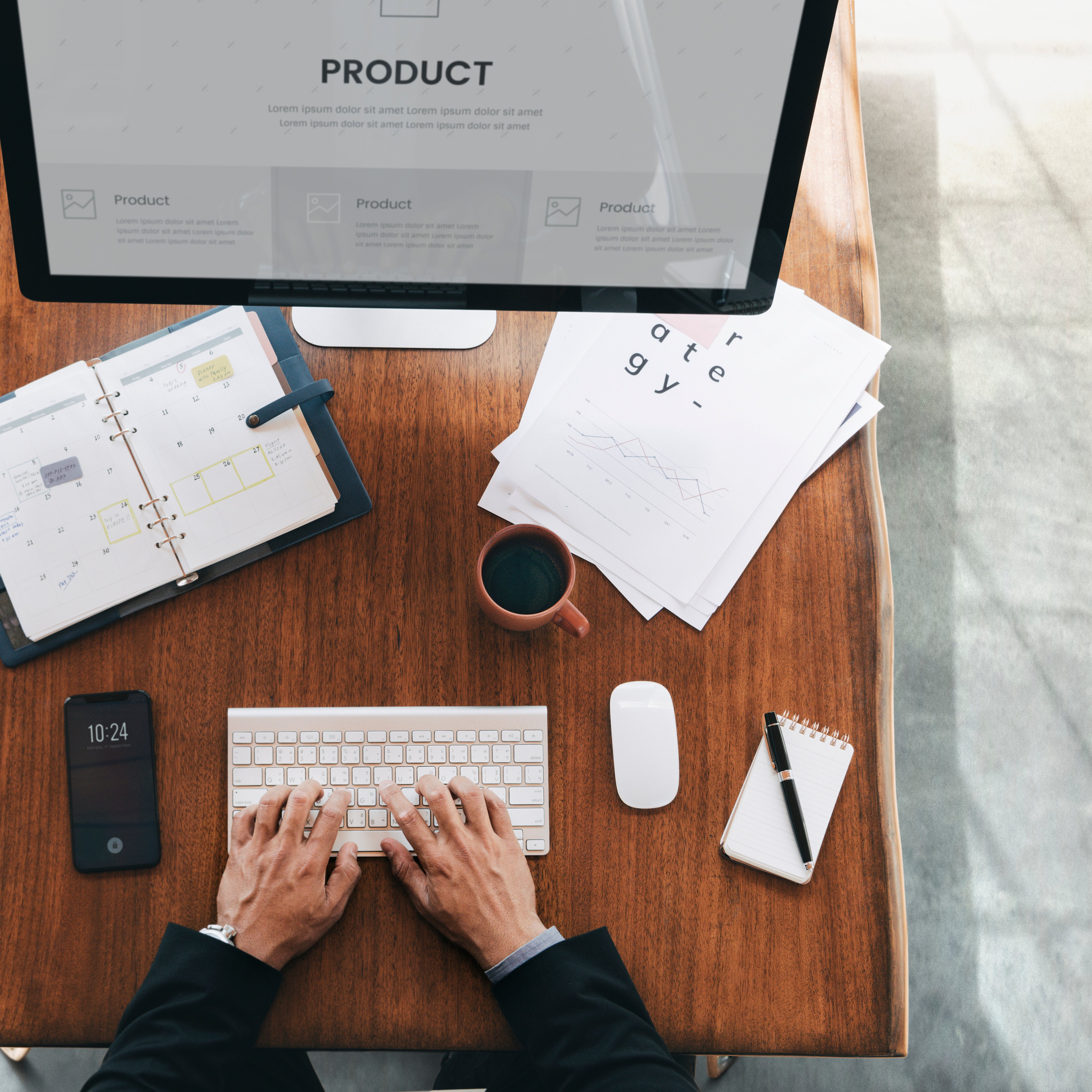 Overhead view of a desk with a computer displaying a product strategy page, open notebook, coffee cup, smartphone, and documents.