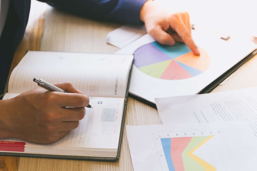 Person analysing charts and graphs at a desk, writing in a planner with a pen, indicating focus on data and planning. 