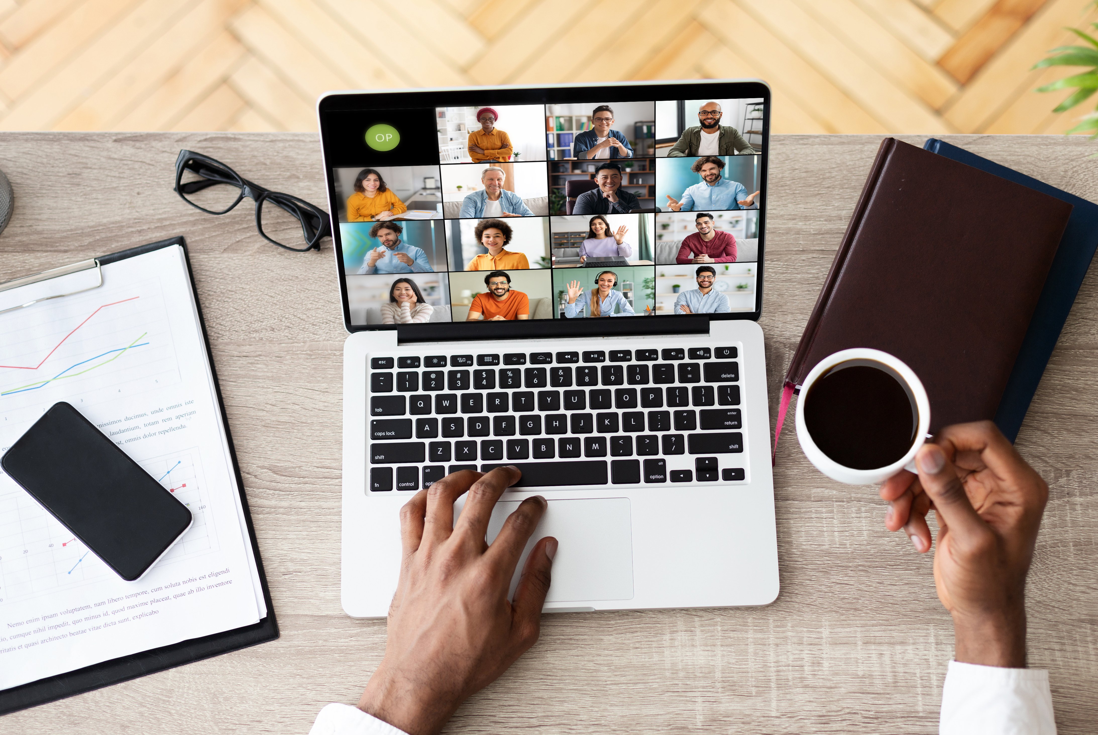 Person attending a video conference on a laptop while holding a cup of coffee, with documents and a phone on the desk. (1)