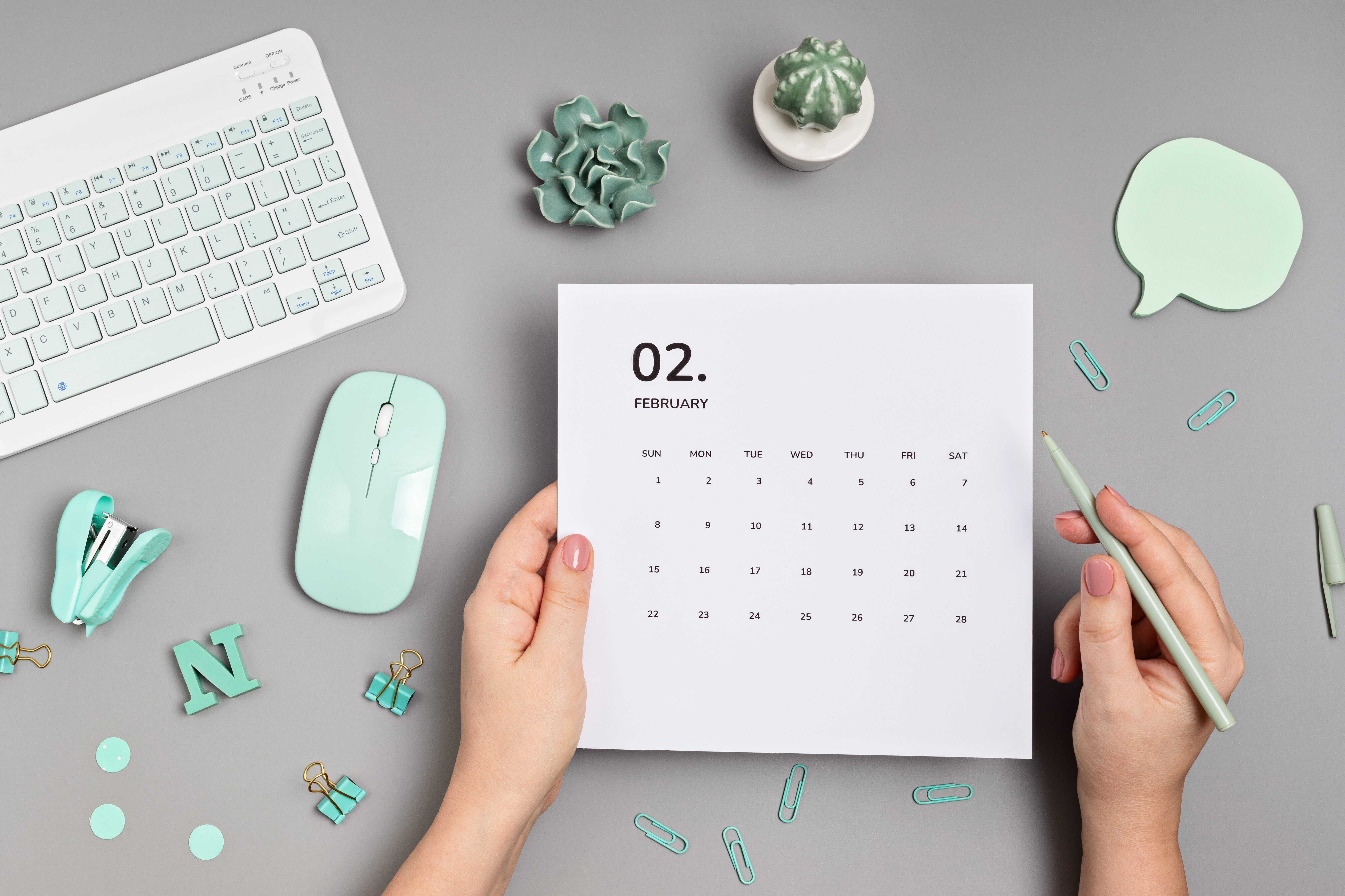 Person holding a February calendar on a desk, surrounded by mint green office supplies, a keyboard, and a small plant