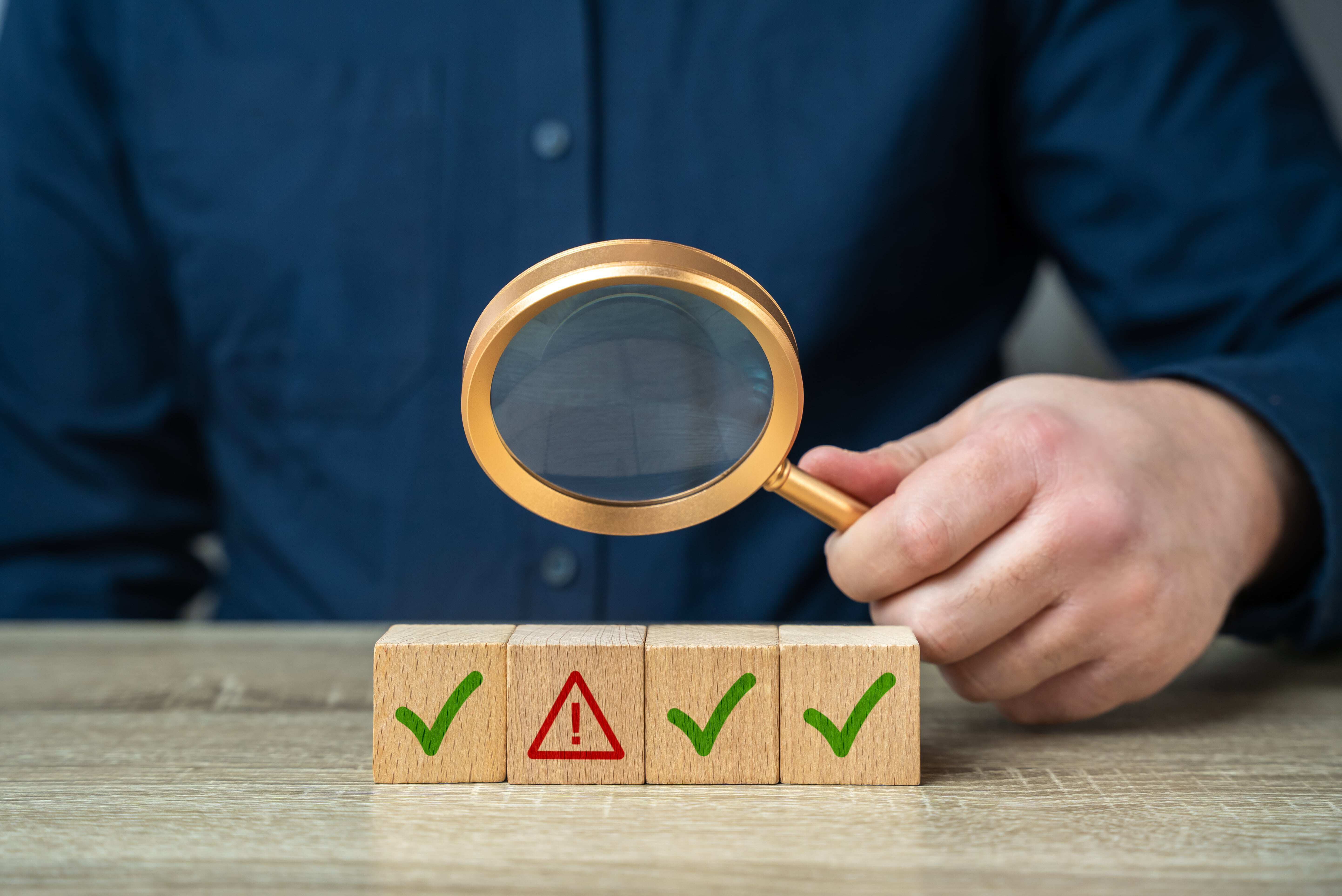 Person holding a magnifying glass over four wooden blocks on a table, with three green check marks and one red warning symbol.-min